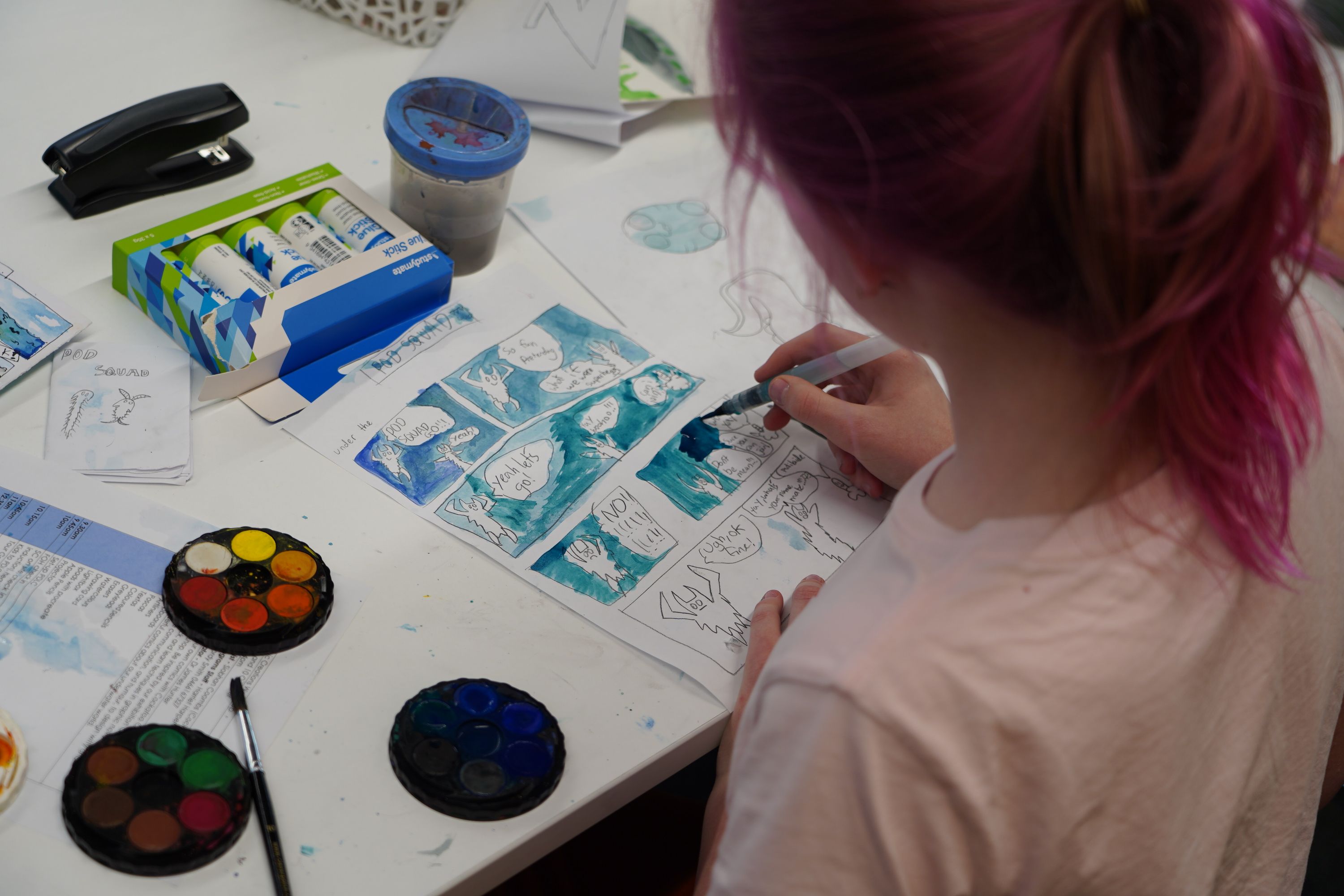 photo over the shoulder of a girl with pink hair in a ponytail. She is painting a comicstrip with watercolour paints.
