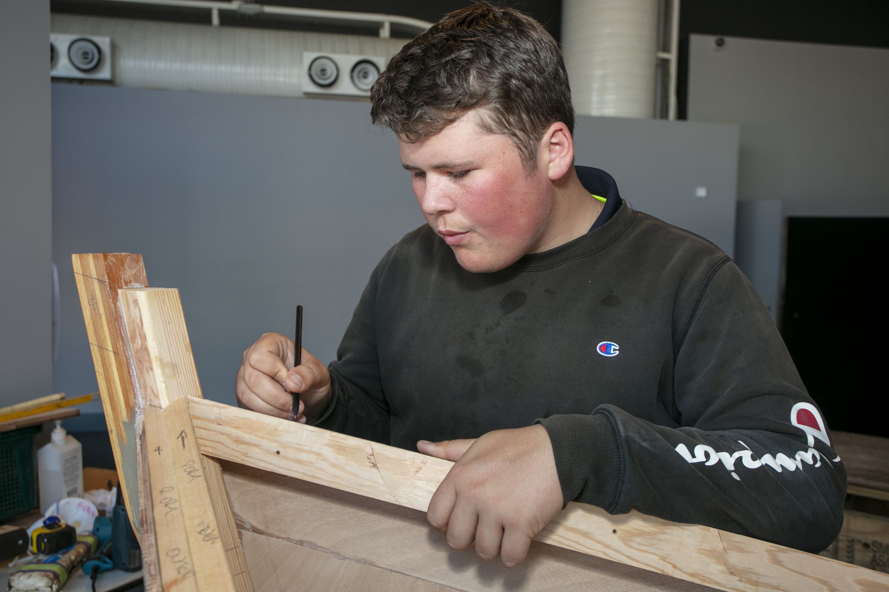 A teen boy wearing a black jumper is using a pencil to assist with building a wooden boat