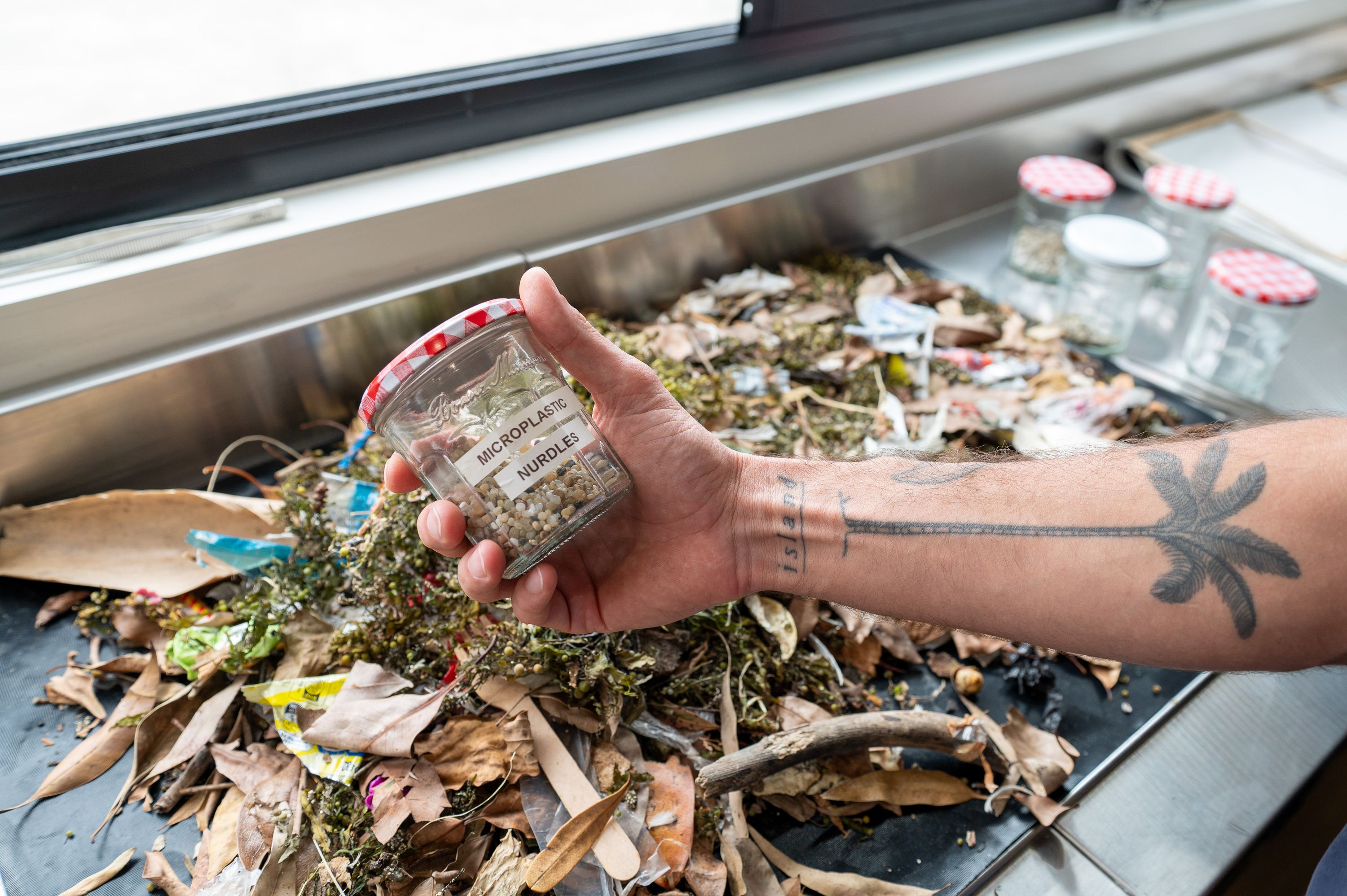 Photograph of a hand holding a jam jar containing small bits of plastic above a table with rubbish and leaf litter.