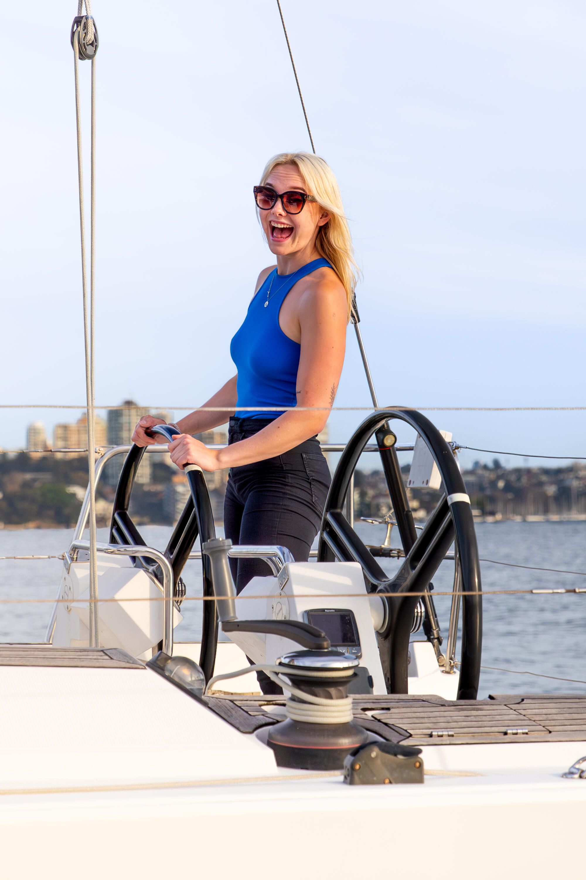 A woman standing with hands on the wheel, with blonde hair, wearing a blue singlet top and sunglasses.  