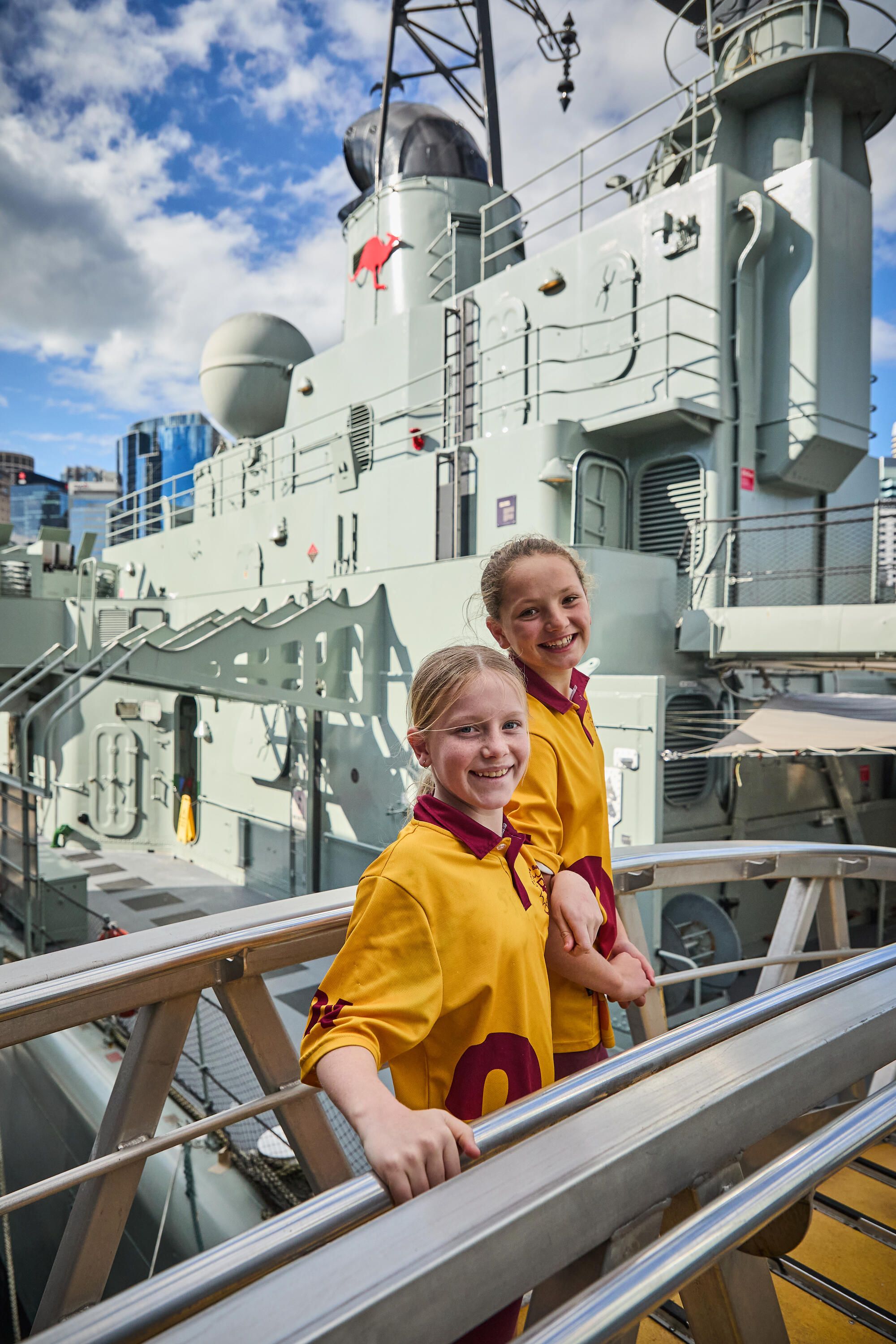 Two girls in a yellow and red school uniform on a gangway with the navy vessel vampire behind them.