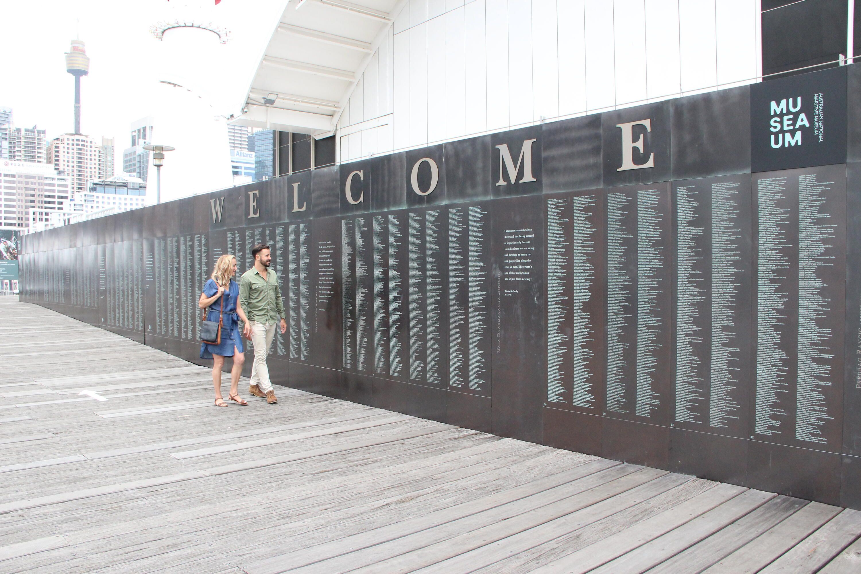 Photo showing a couple walking outside the museum past the Welcome Wall name panels.