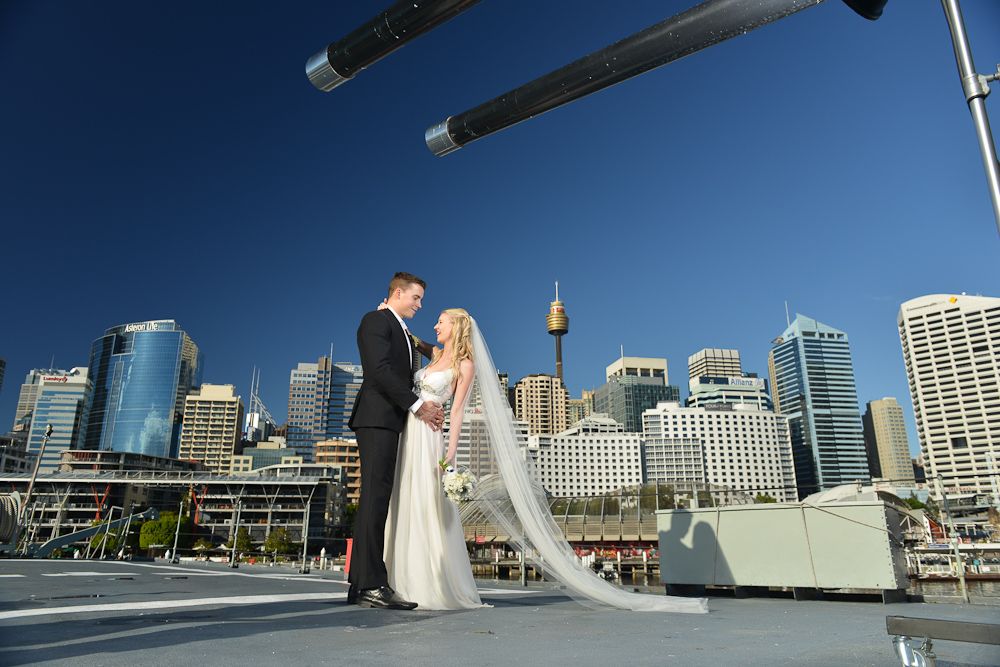 Photo of a bride and groom together on the deck of a ship with the Sydney skyline ad a blue sky behind them.