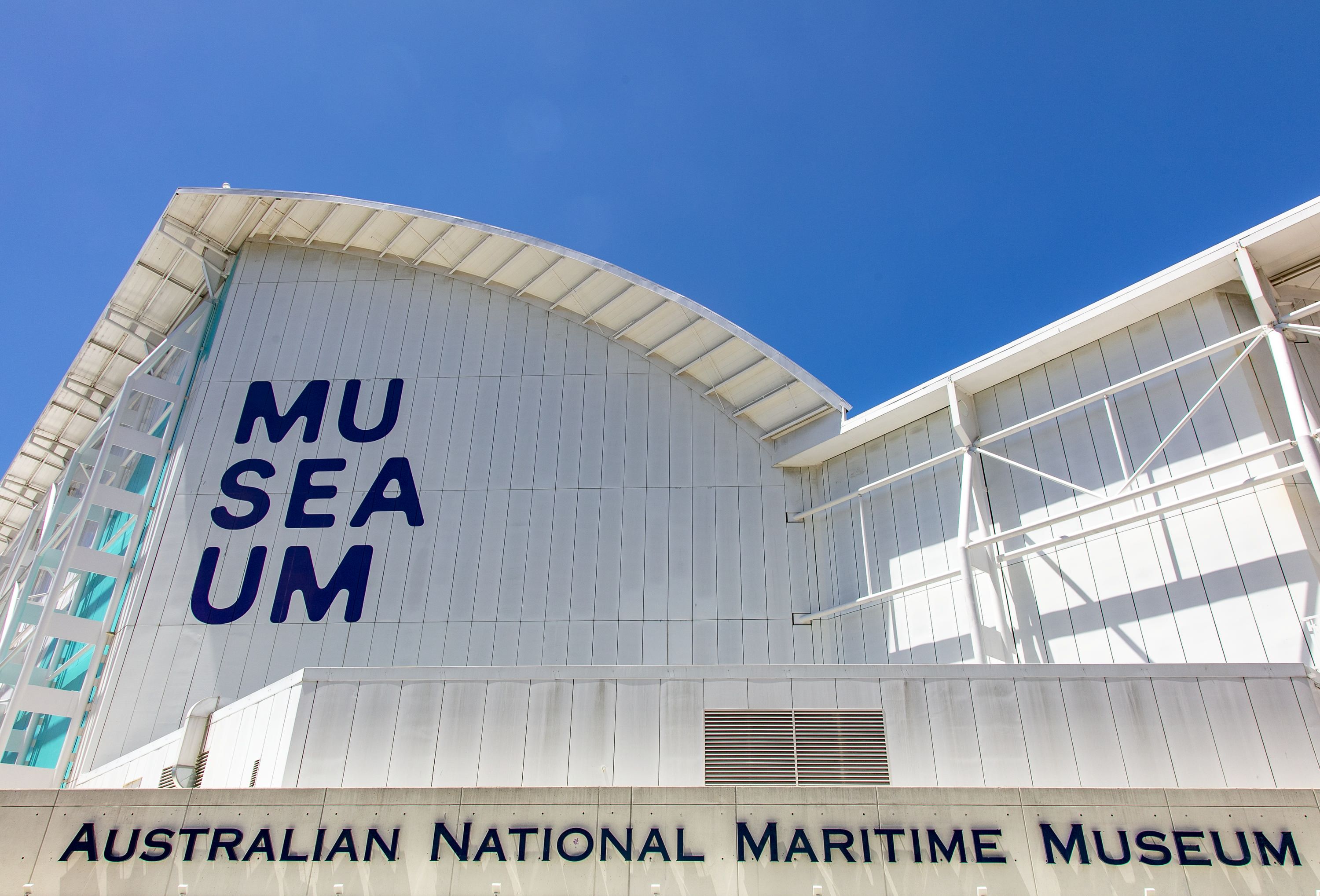 Photograph of the white, museum building with curved roofline, and the museum logo painted on the wall. 
