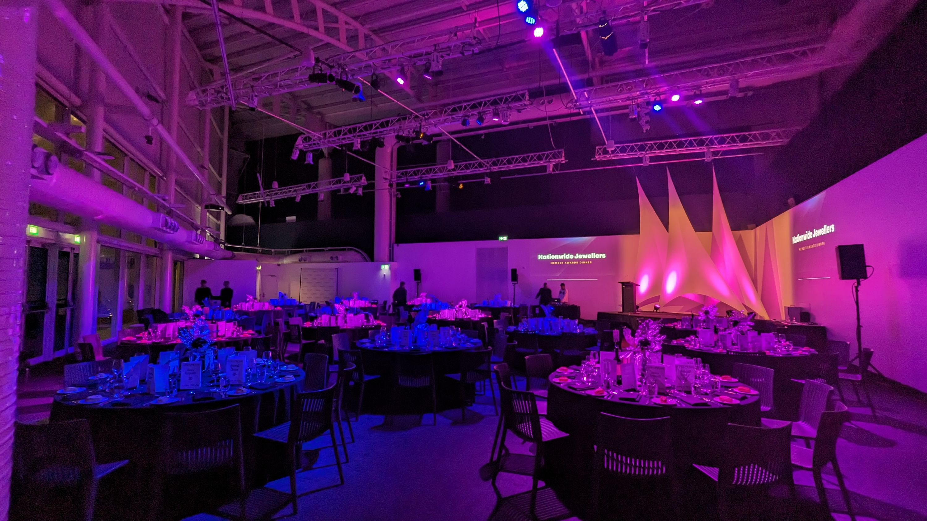 Inside an event space at night. The room is lit in pink, with round tables and a stage in the corner.