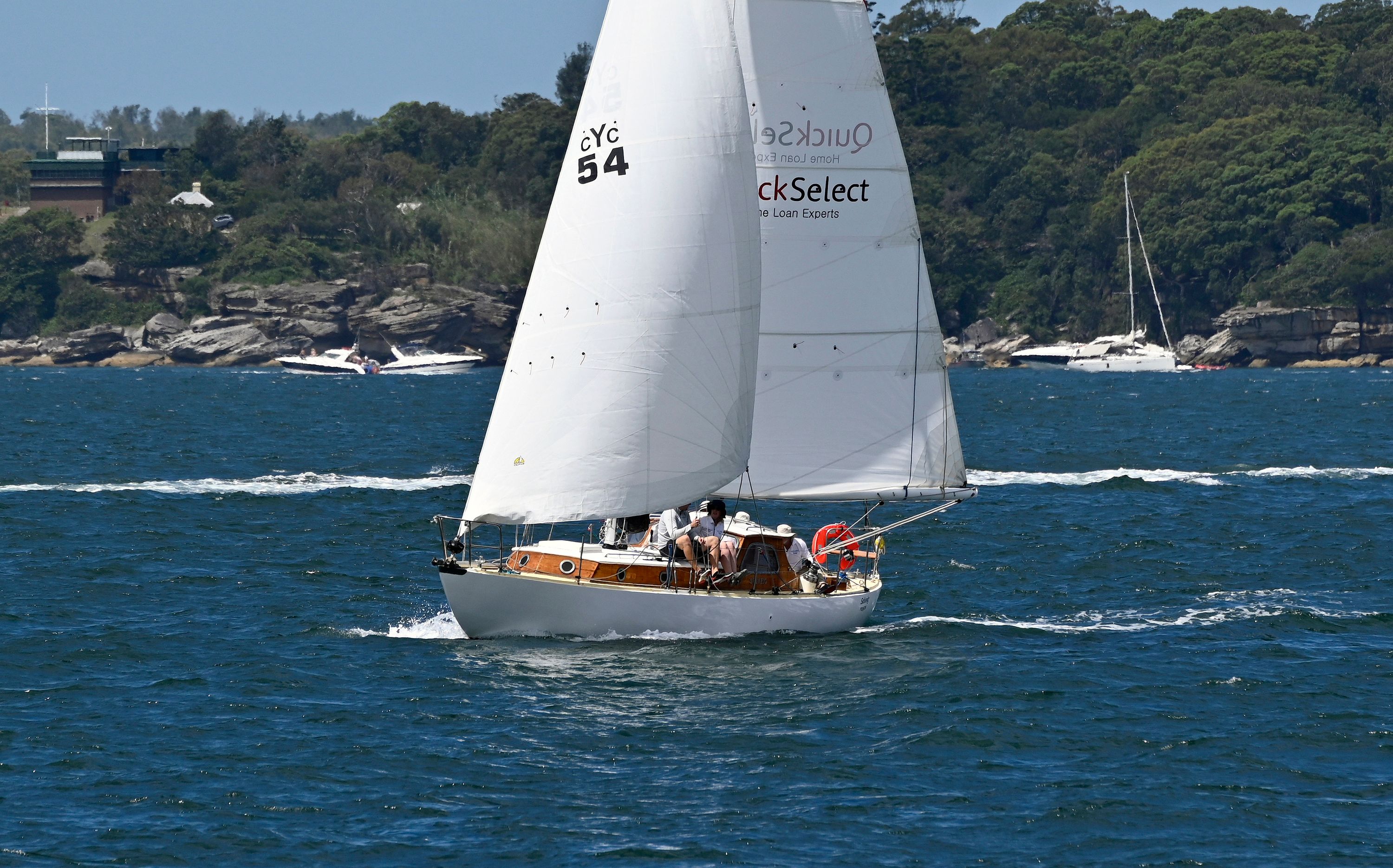 A photo of a small boat with 2 large triangular sails filled with wind, sailing on a dark blue water, with green foliage and a clear sky behind it. 