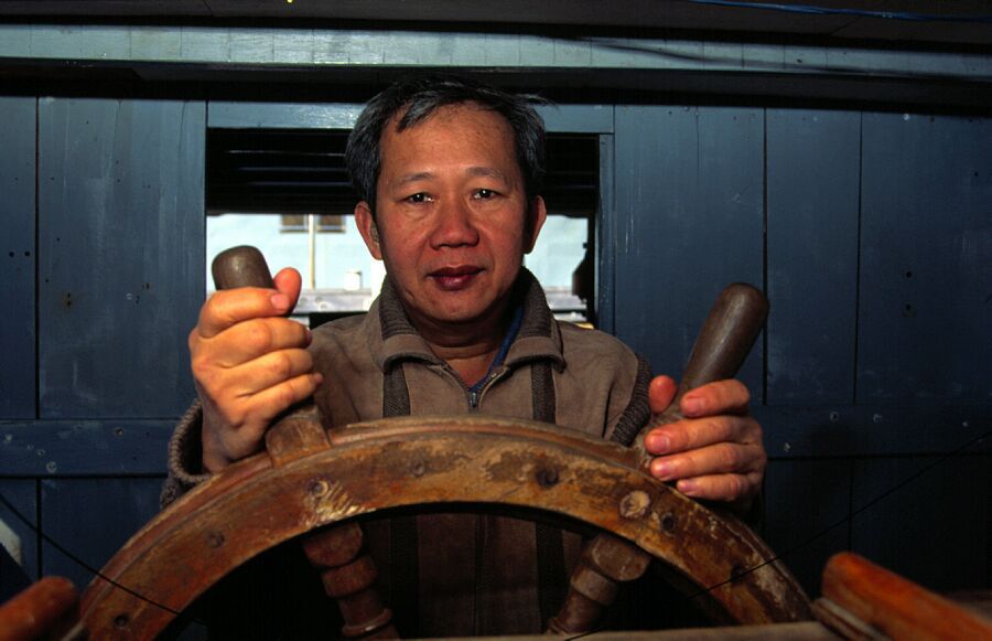 Photo of an older Vietnamese man standing at a ship's wheel in the cabin of a wooden boat.
