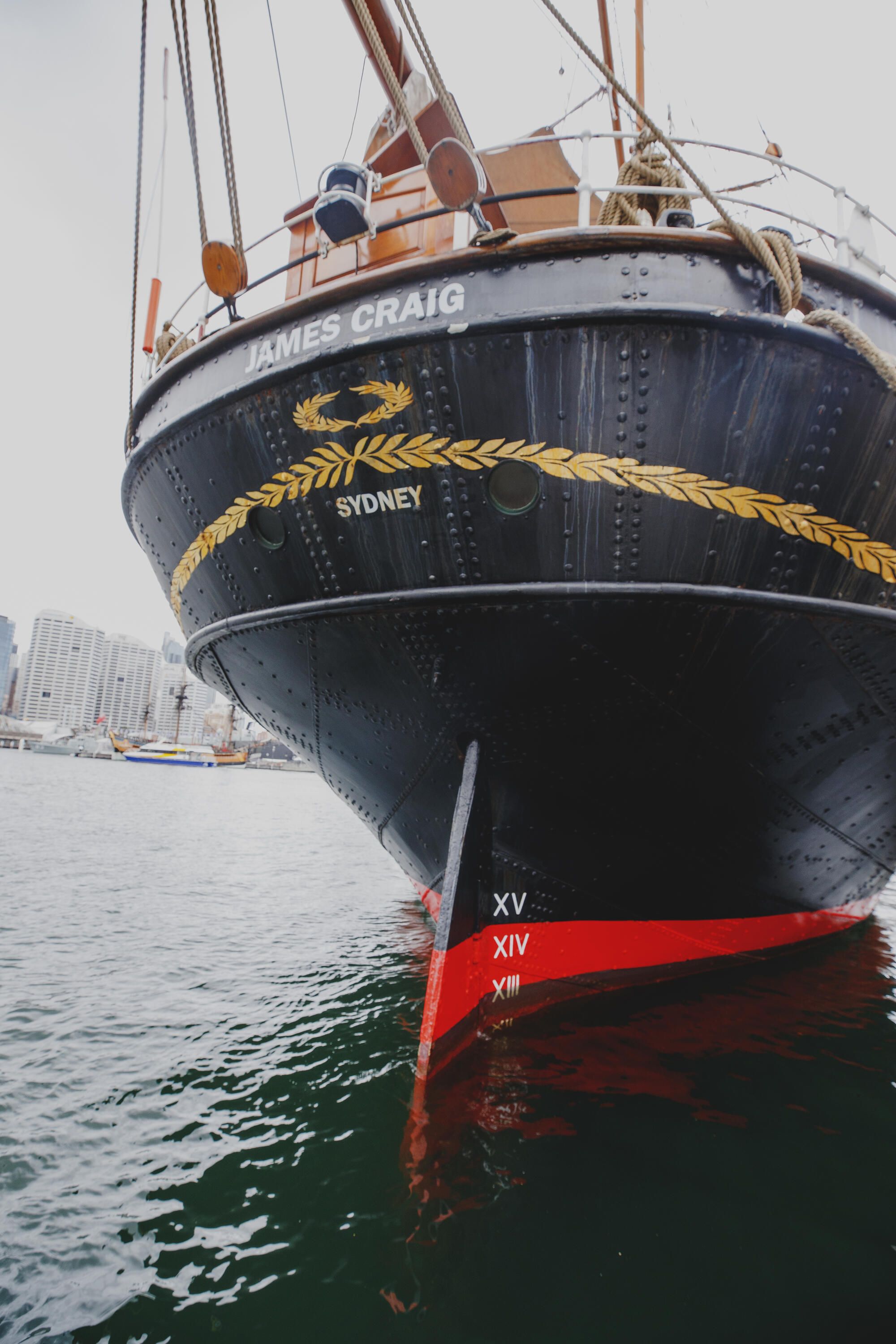 Photo showing the rear of a tall ship's hull, painted black with red at the waterline and gold details. "James Craig" and "Sydney" are painted in white.