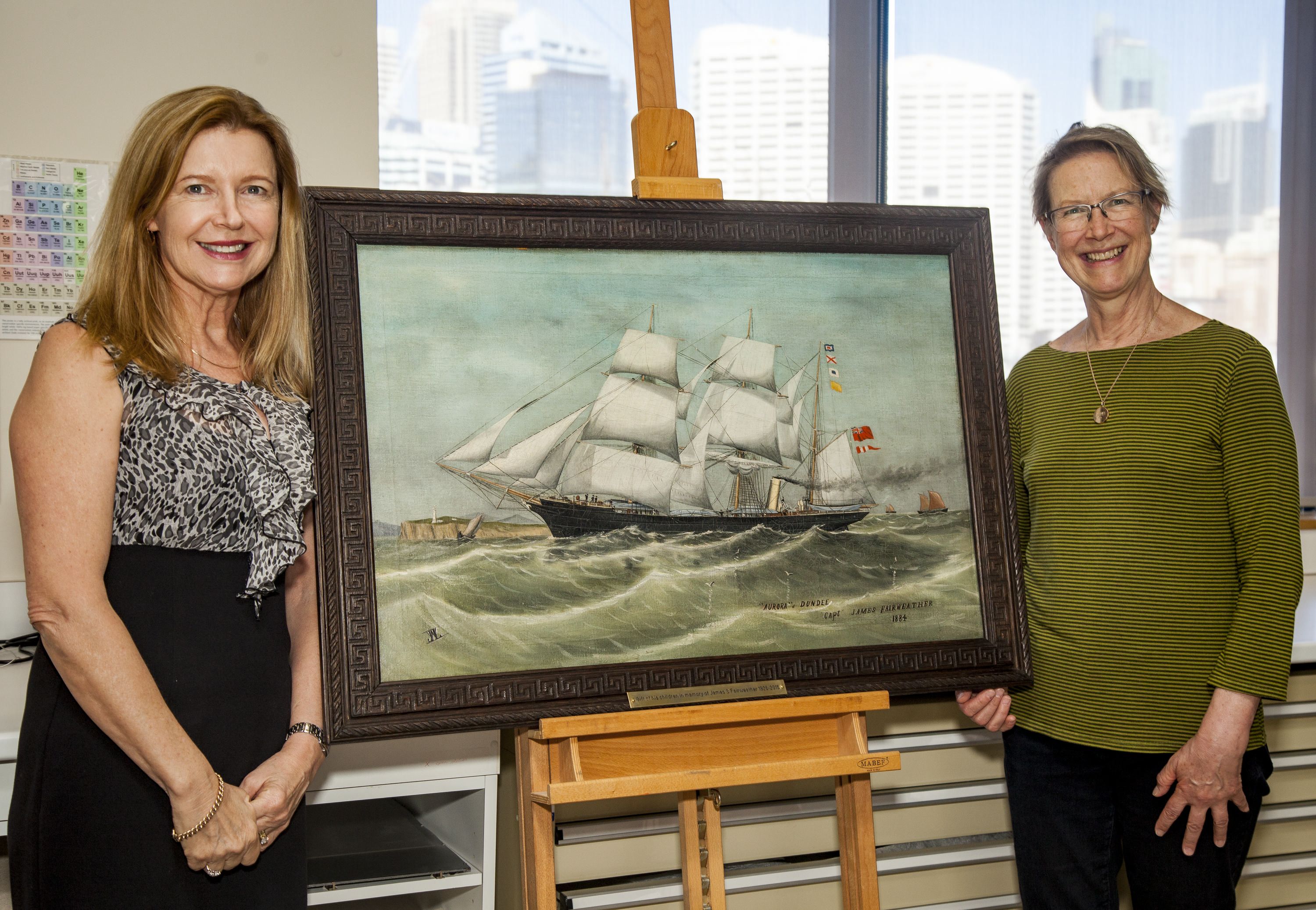 Photograph depicting two smiling women standing either side of a framed painting of a sailing ship with white sails. 
