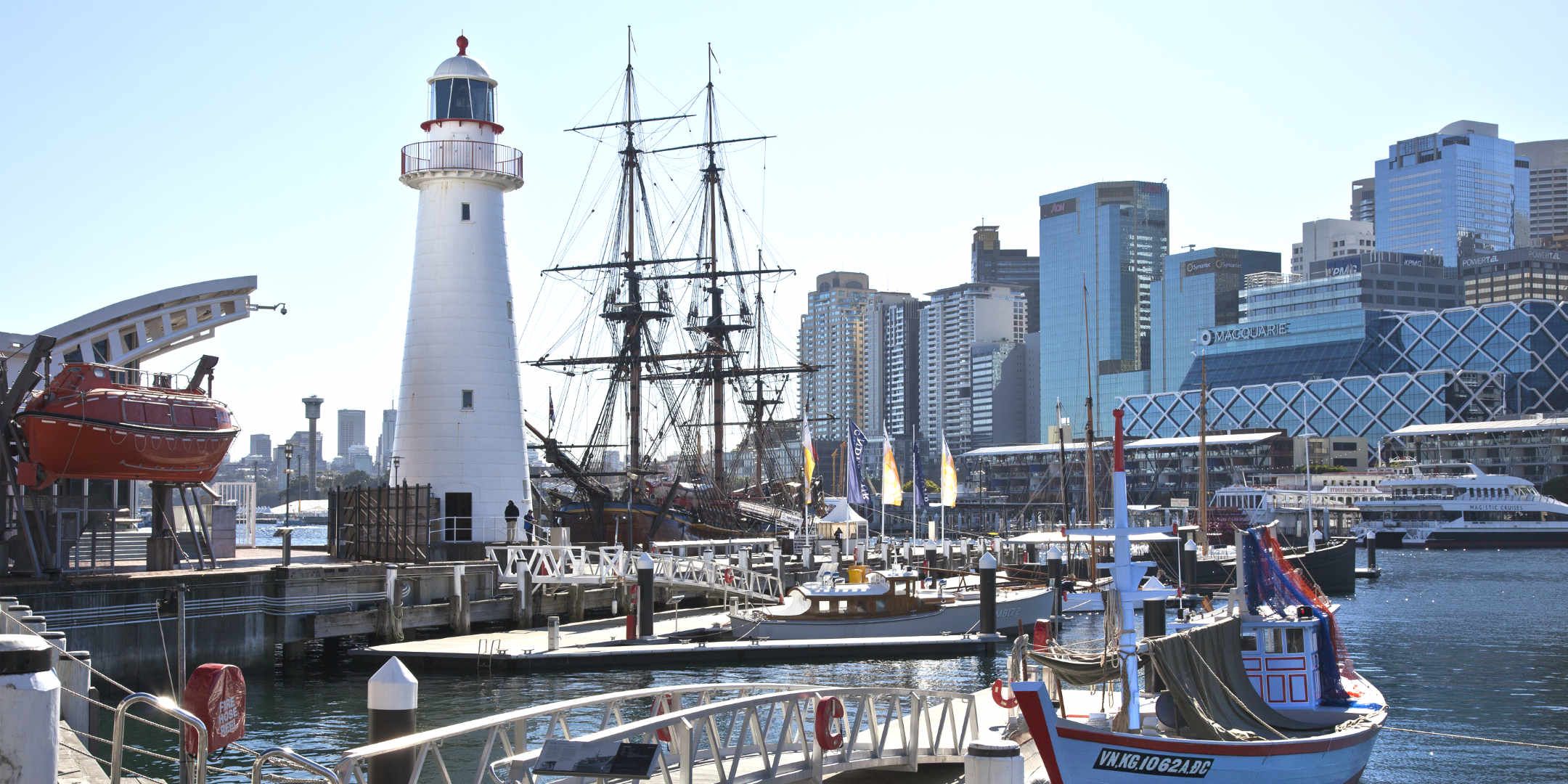 Photo showing a view of the harbour featuring museum vessels moored at the wharves and a white lighthouse.