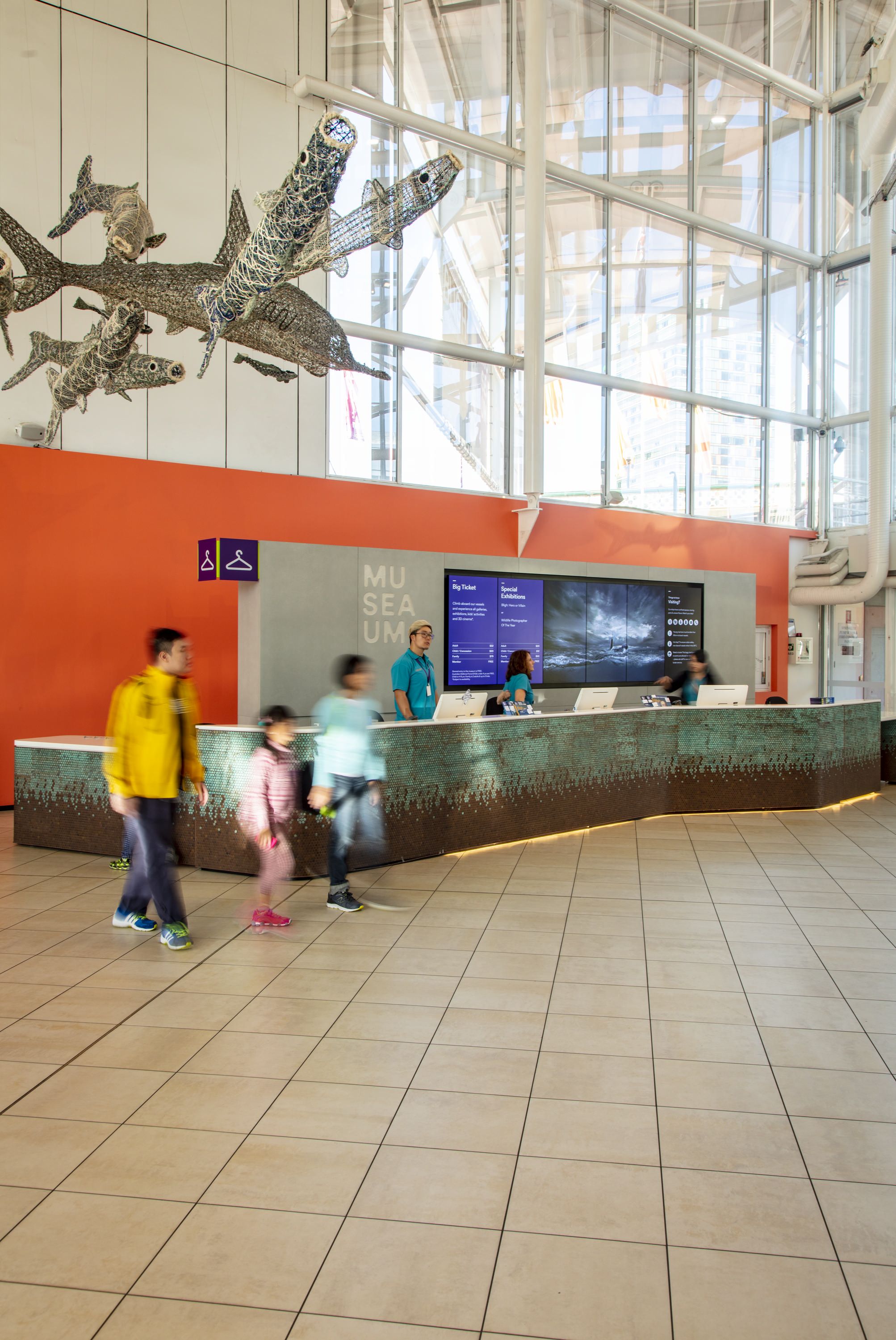 Photo showing the museum foyer near the front desk. Bury figures of a family are captured in movement. The Ghost Net sculptures appear overhead. 