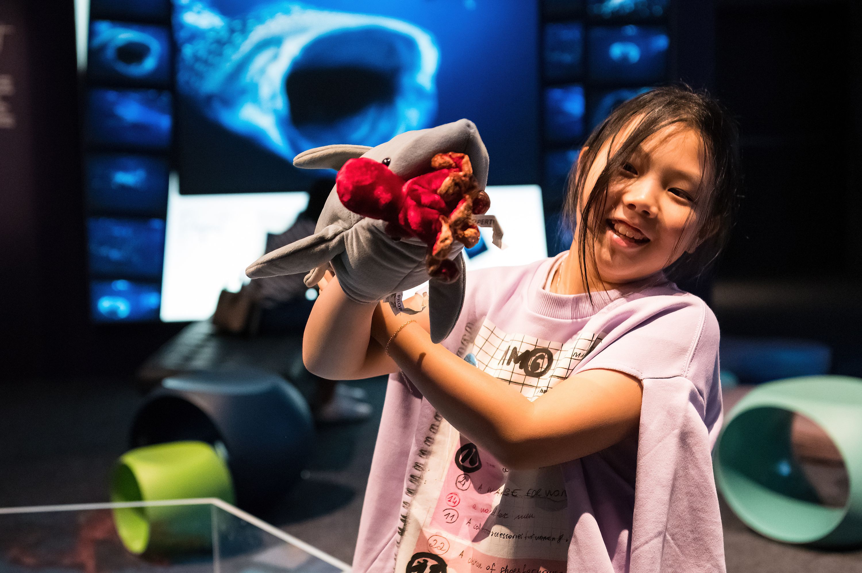 Girl with a shark puppet performing for the camera. Behind her is a photo of a whaleshark