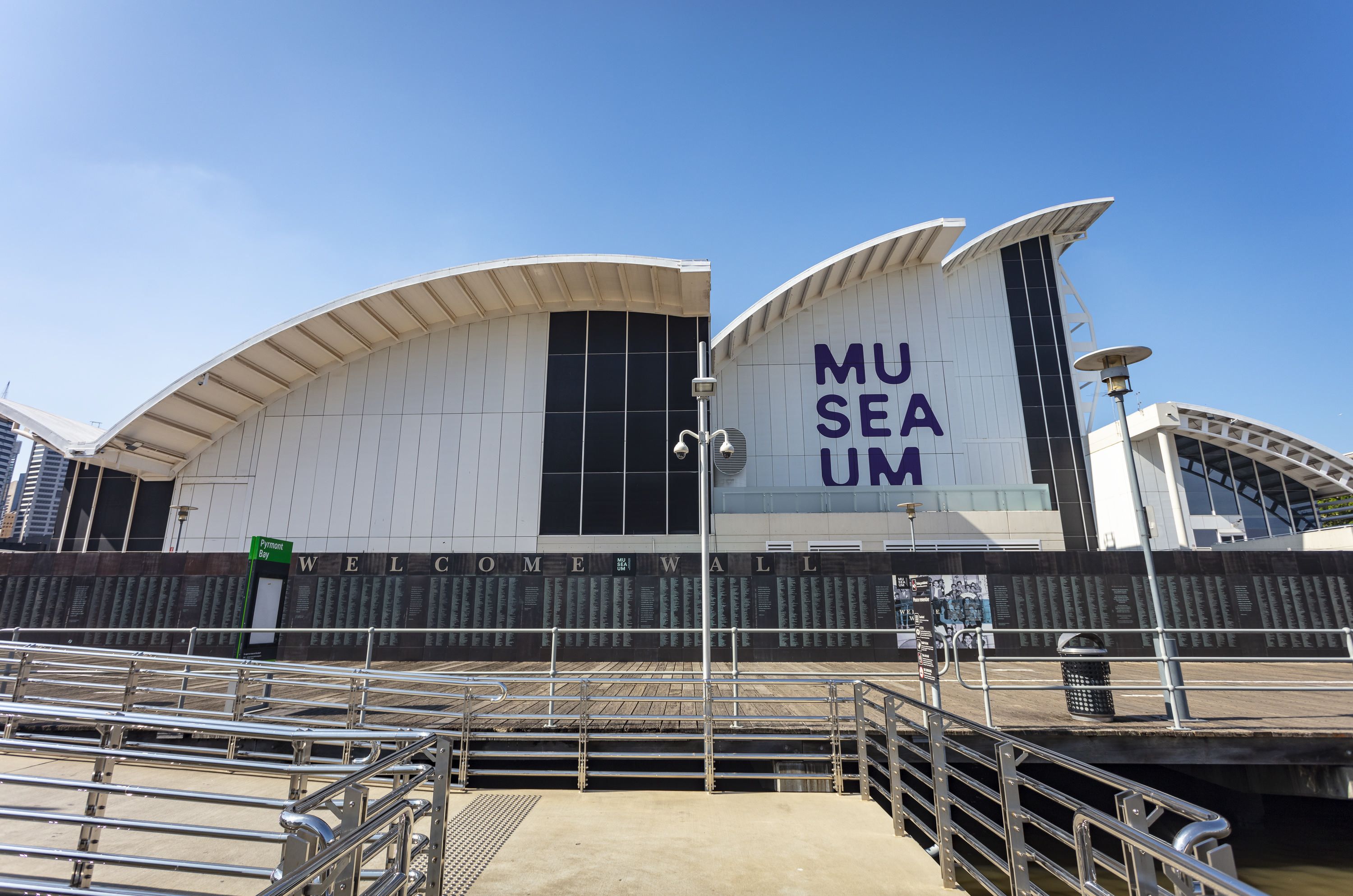 Photo of the museum building behind the welcome wall, a bronze wall with engraved names.  The museum logo is on the white wall, and a blue sky behind. 