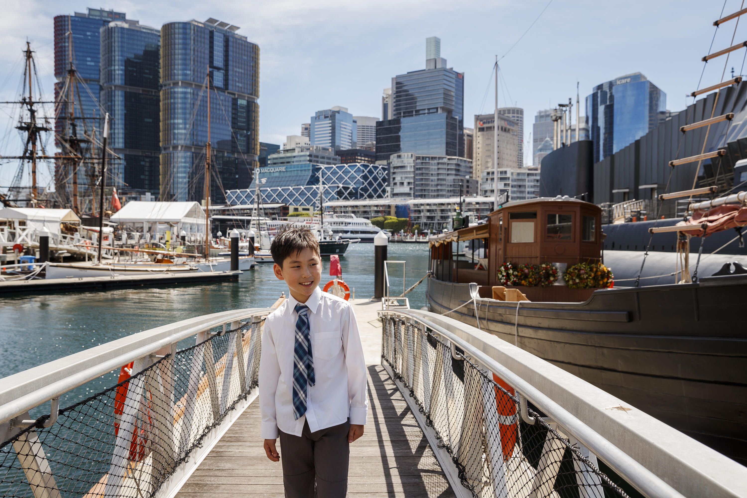 Photo of a boy on a walkway next to a wooden boat, with the Darling harbour and the Sydney skyline in the background.