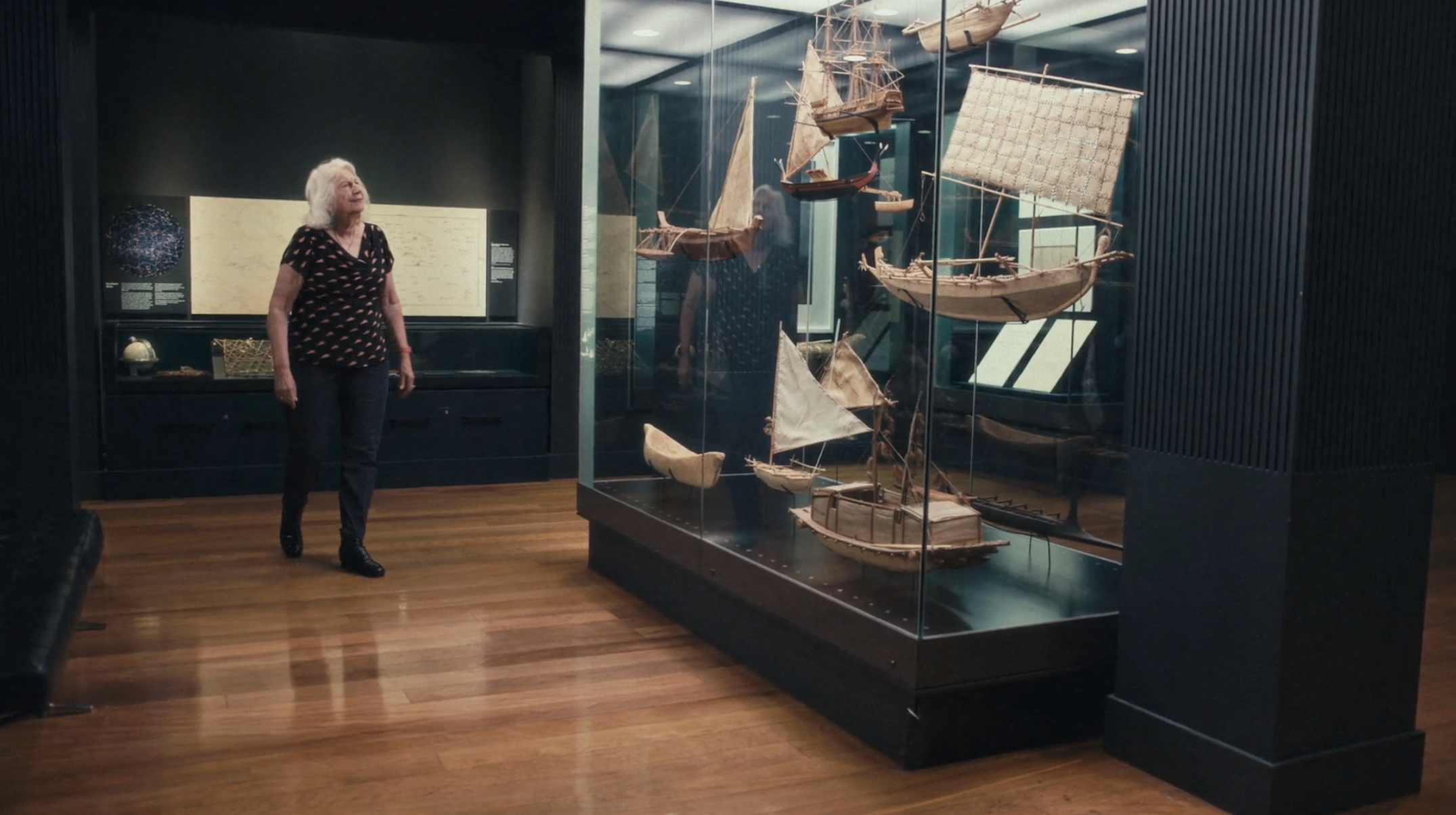 Photo of a woman walking through a museum gallery looking at a display of model ships. 