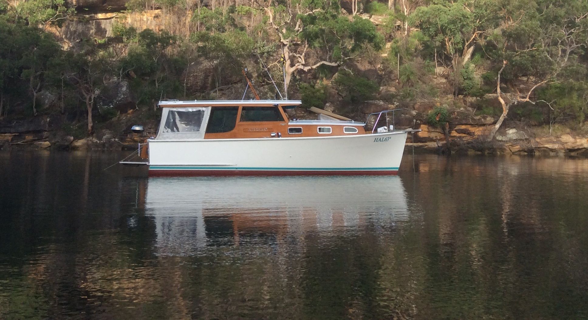 Photo of a boat with a white hull and wooden features, still on the water with green foliage behind it. You can see the reflection of the boat in the still water.