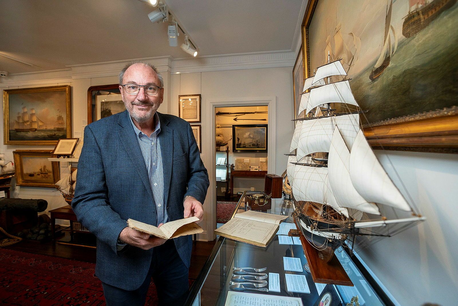 Photo of an older man in a suit jacket holding a book and looking at a model of a sailing ship.