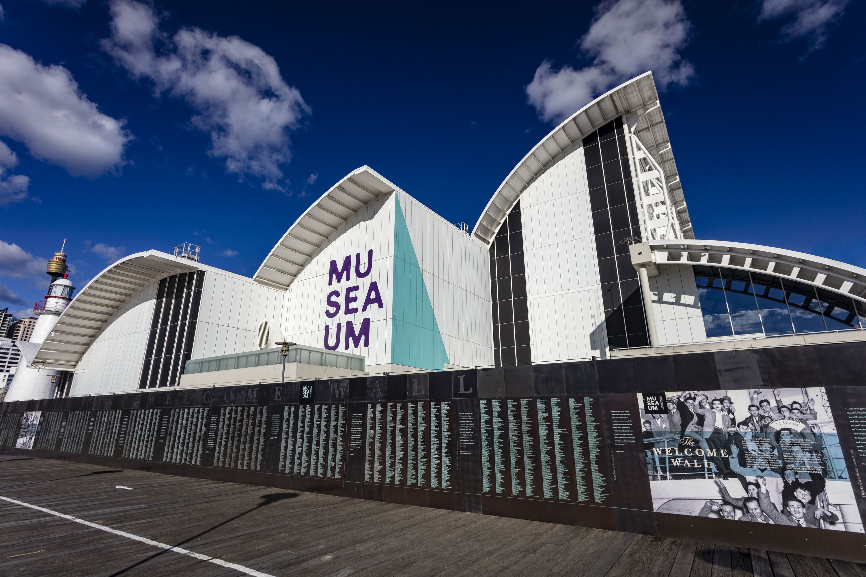 Photo showing a dark metal wall with green engravings. It is in front of a large white building with the museum logo.  