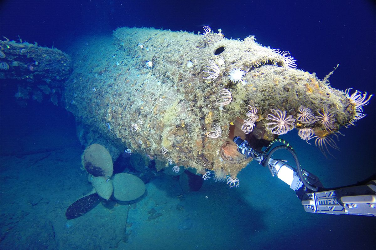 Photo taken underwater depicting the rear of a submarine that has been sunk.it is covered with marine life, and a robotic arm can be seen at the right of the image.