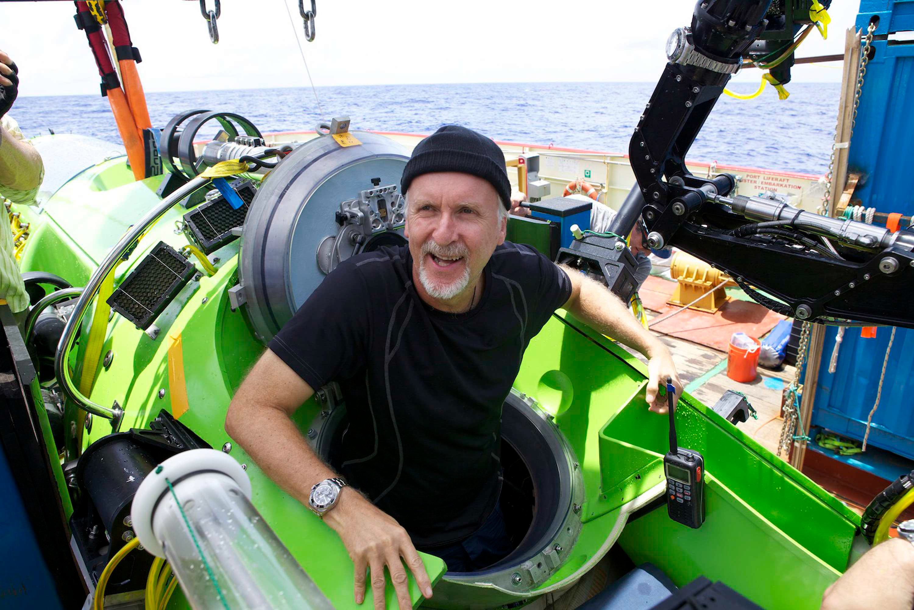 Photo showing a man, James Cameron, wearing a black t-shirt and beanie, comming out of a green submarine.