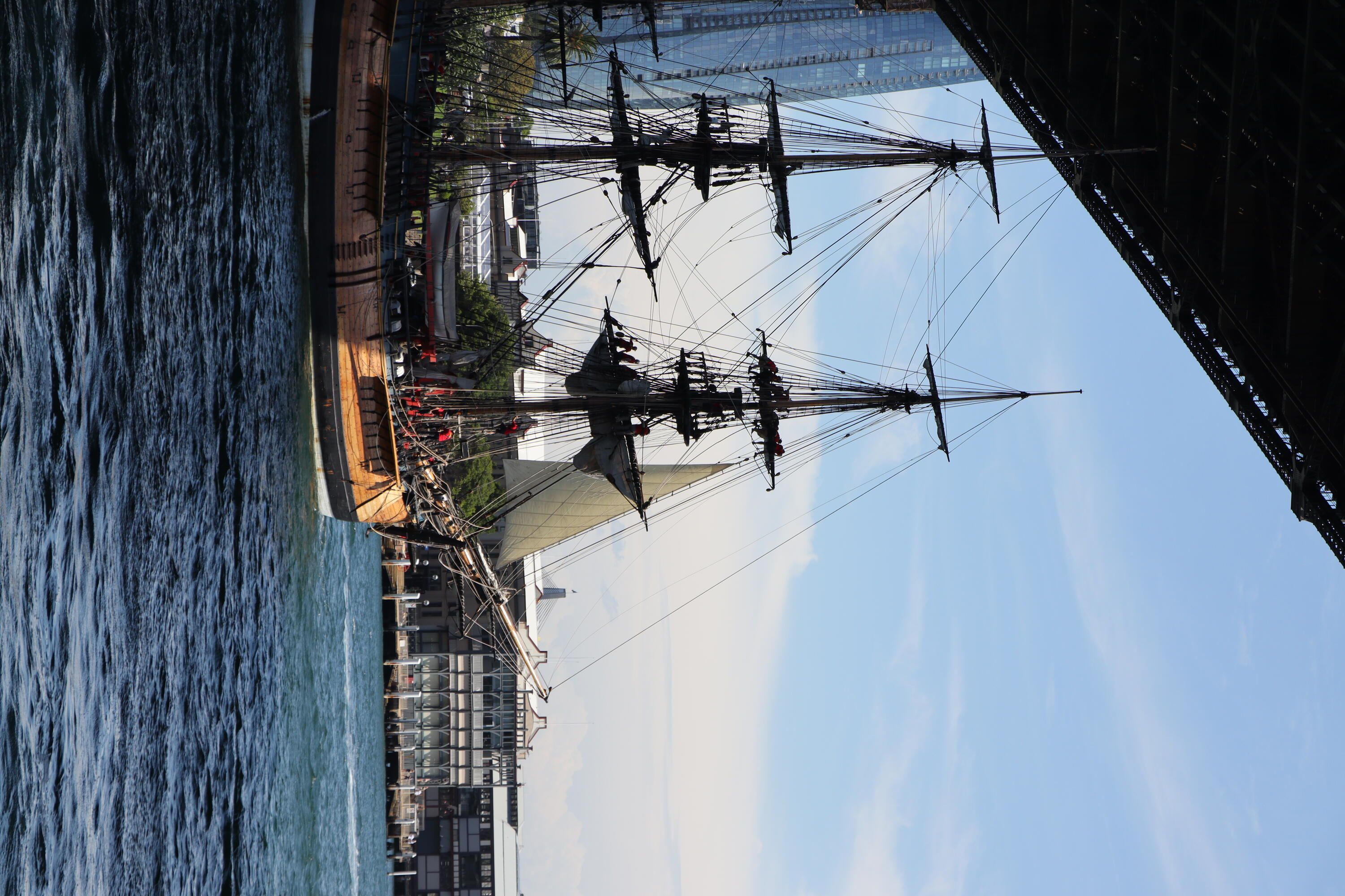 Photo showing a tall ship, Endeavour, sailing under the Sydney Harbour bridge, the front is in sunlight, the back is in shadow. 