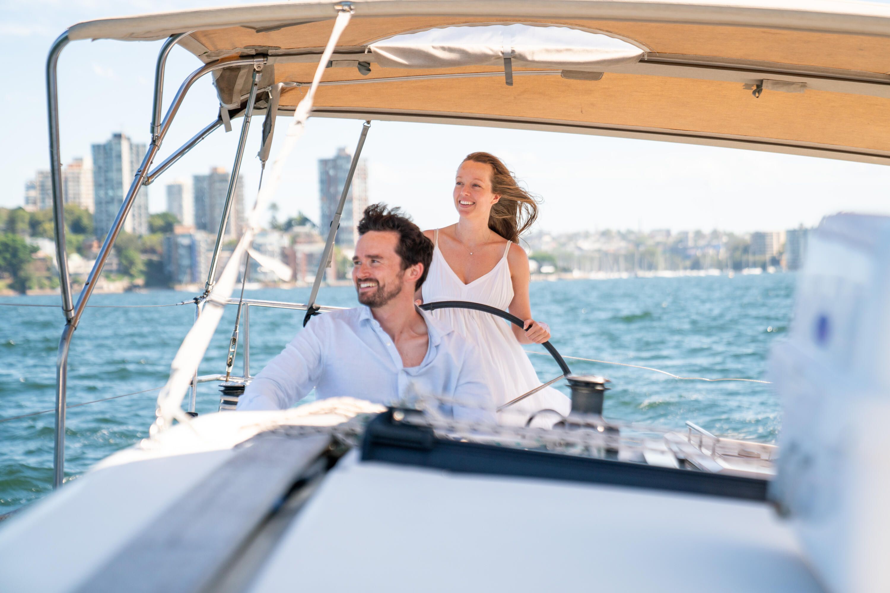 Photo of a man seated and woman wearing a white dress, standing at the wheel of a yacht, with Sydney harbour in the background.