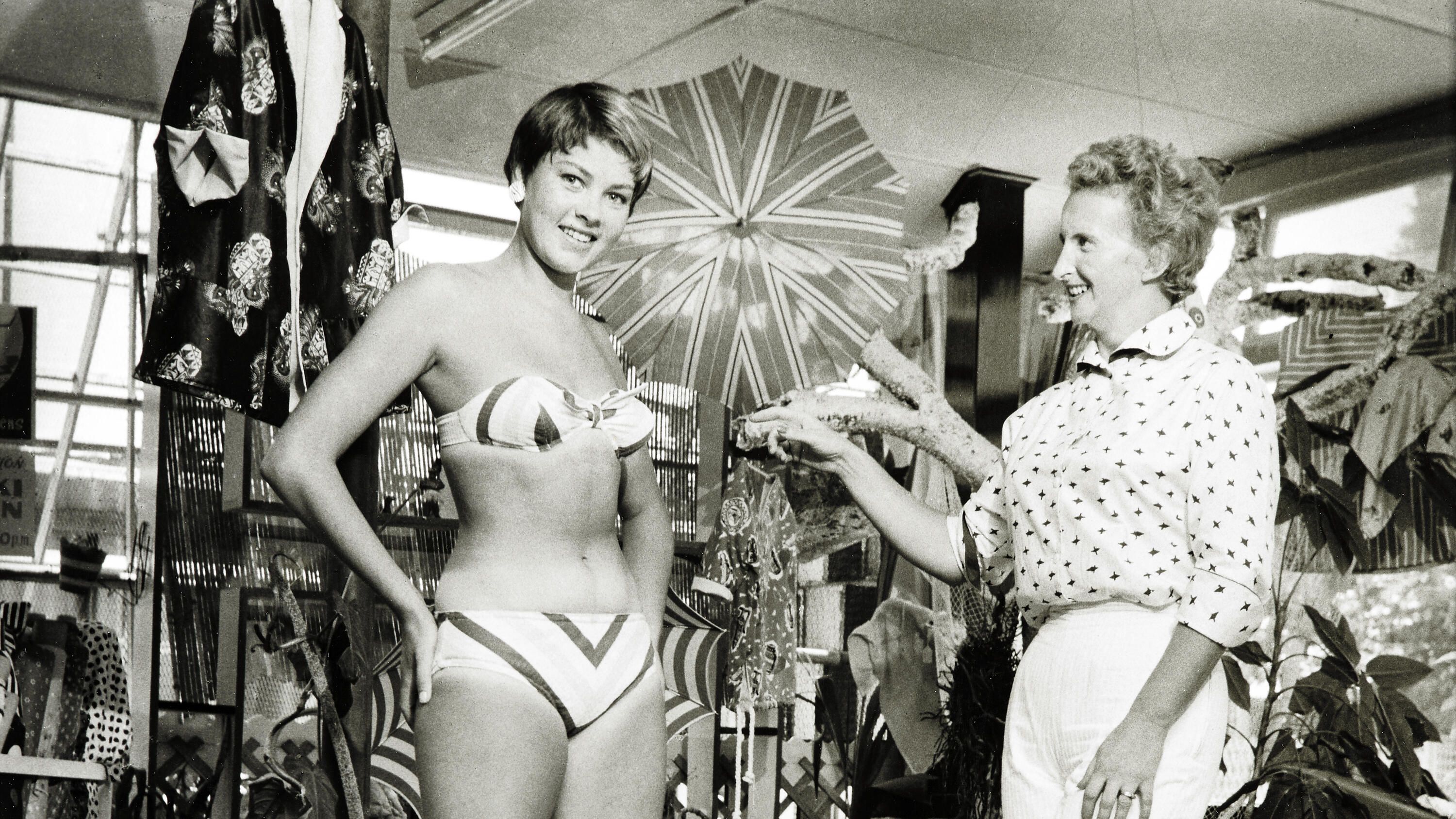This black and white photograph depicting the interior of Paula Stafford's Tog Shop at Broadbeach Gold Coast, Queensland about 1957. Paula is seen standing to the right, gesturing towards her customer, to her left.