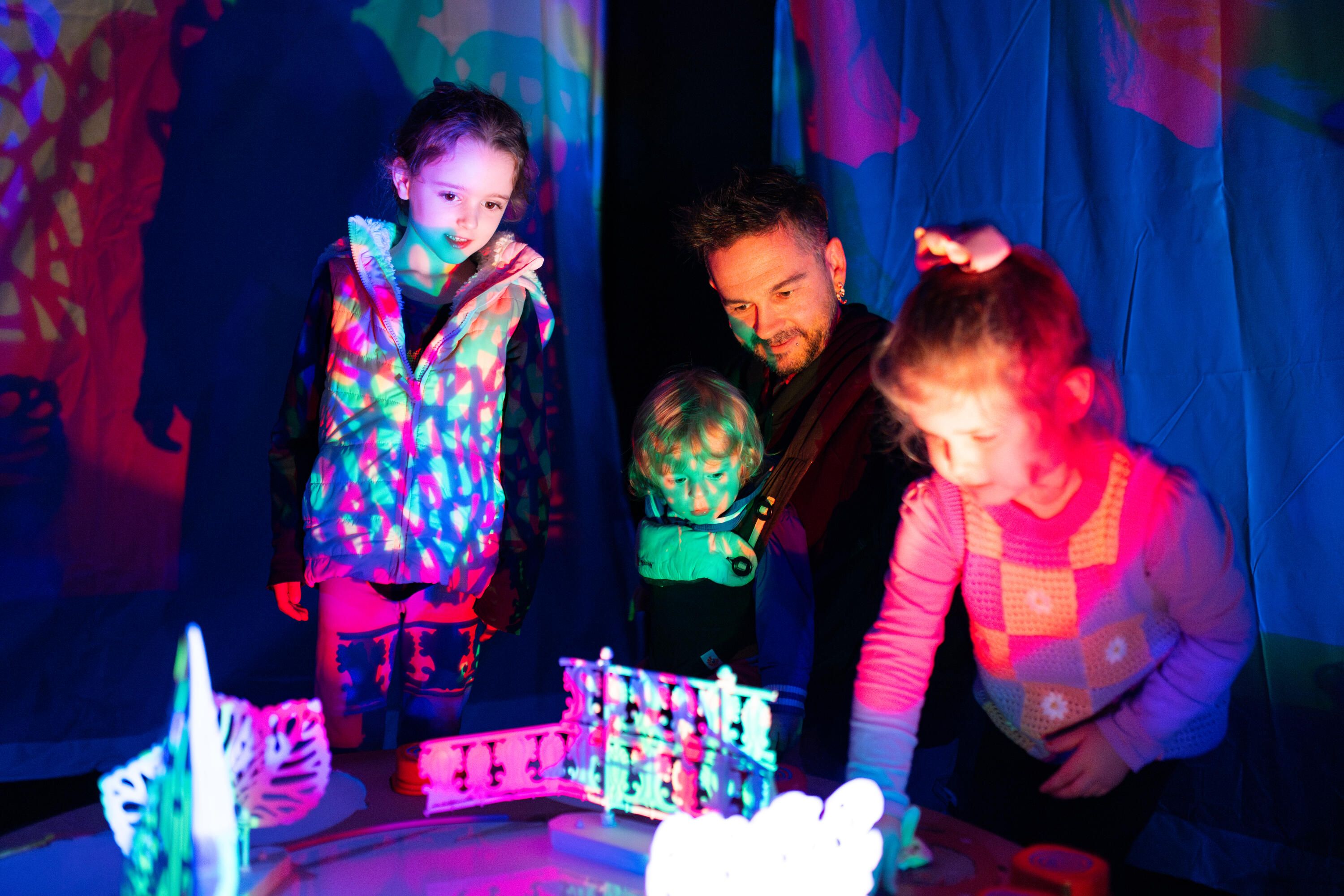 Two children and an adult playing with interactive theatre props in colourful light.