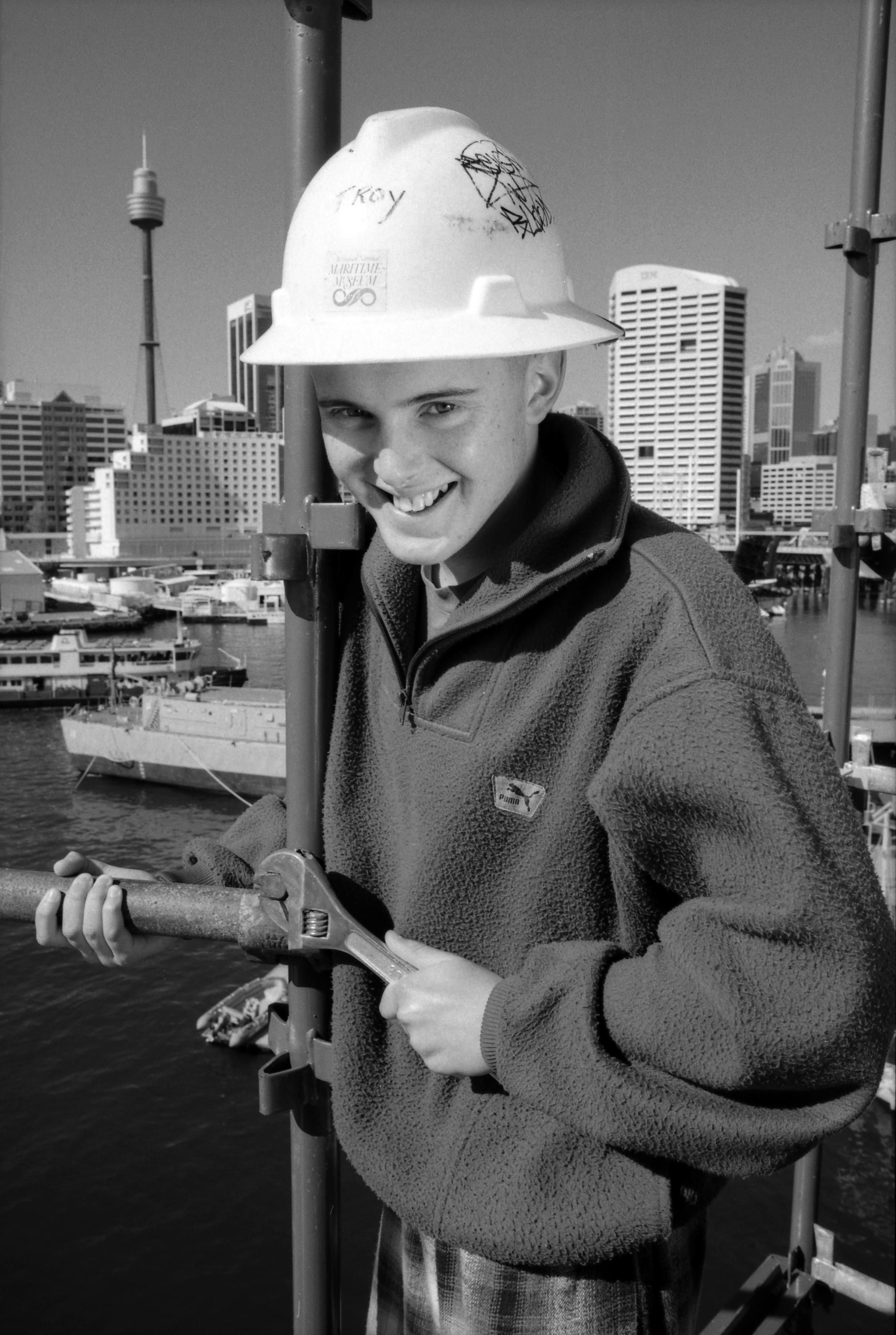 A photo portrait of a young man at work on the project with the city in the background. His hard hat is labelled 'Troy' and he is pictured standing on the scaffolding holding a large wrench