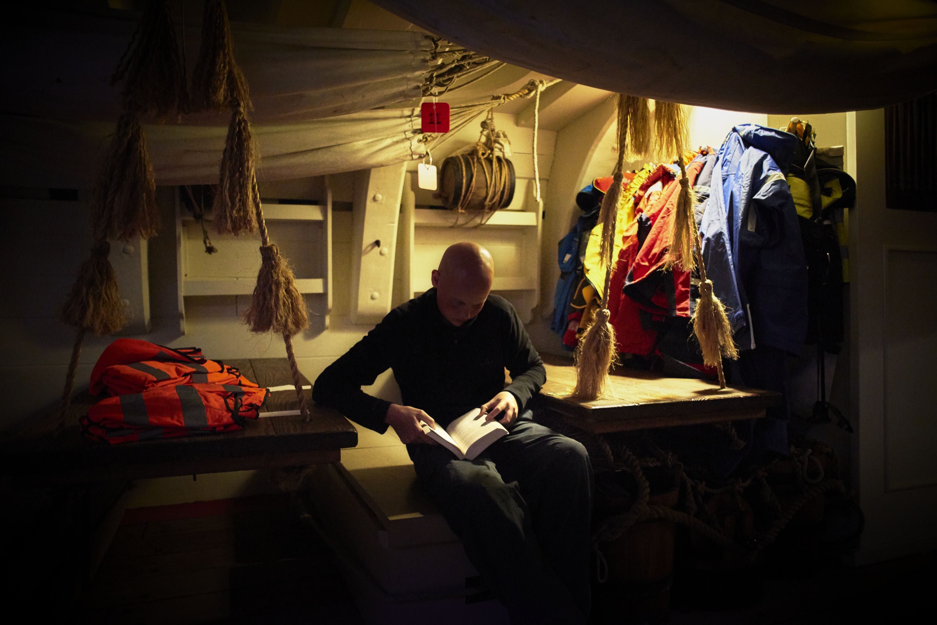 Photo taken of below deck on a tall ship showing a man reading. 