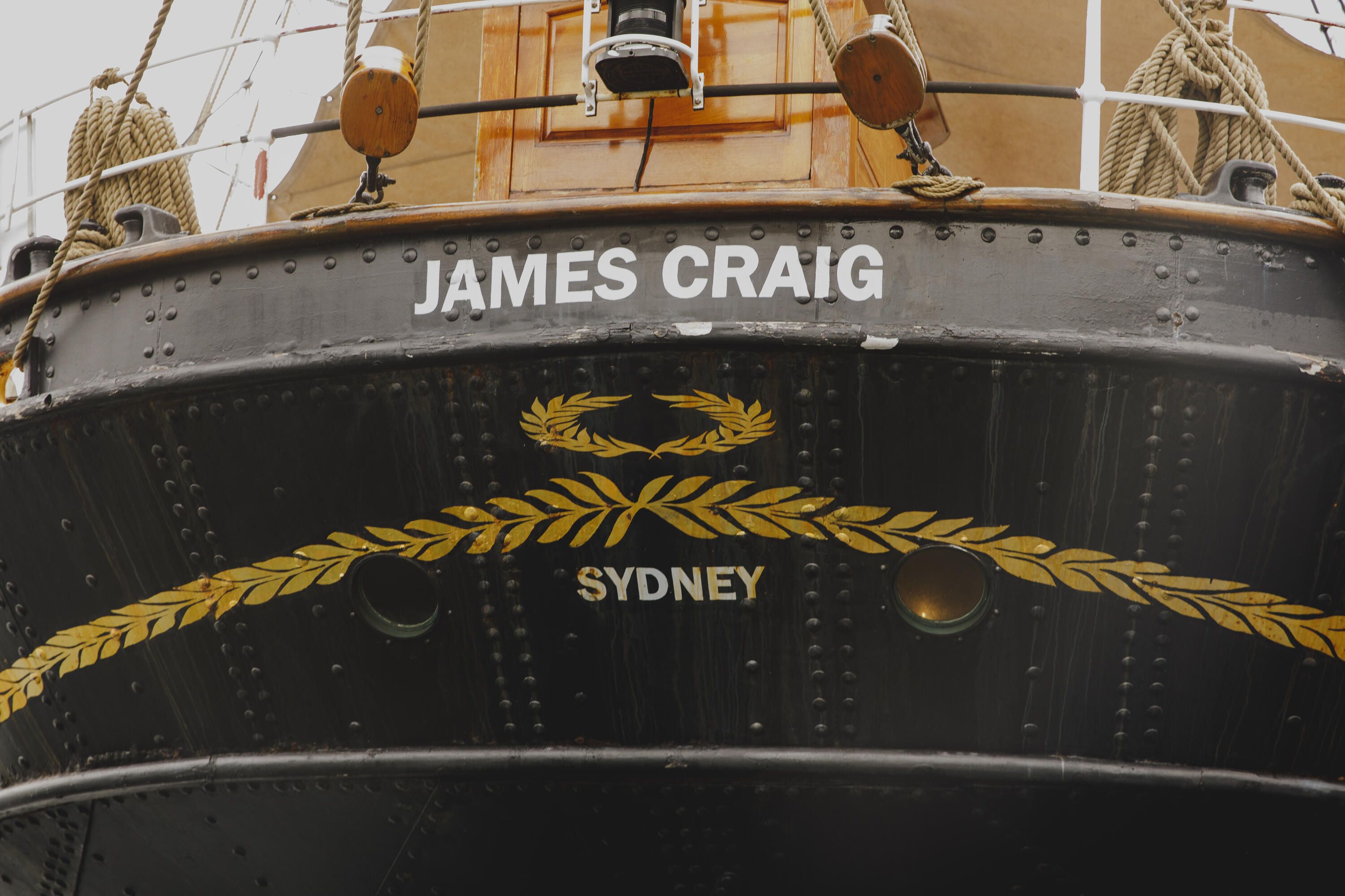 Photograph showing details of the back of a tall ship, featuring the name "James Craig" and word "Sydney" painted in white on the black hull.