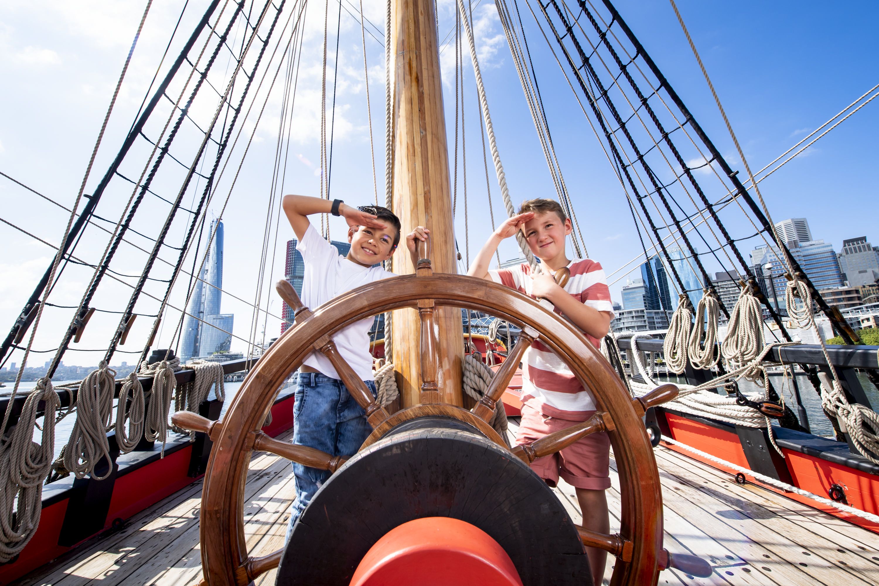 Photo showing two boys on board a tall ship and pose behind the ship's wheel.