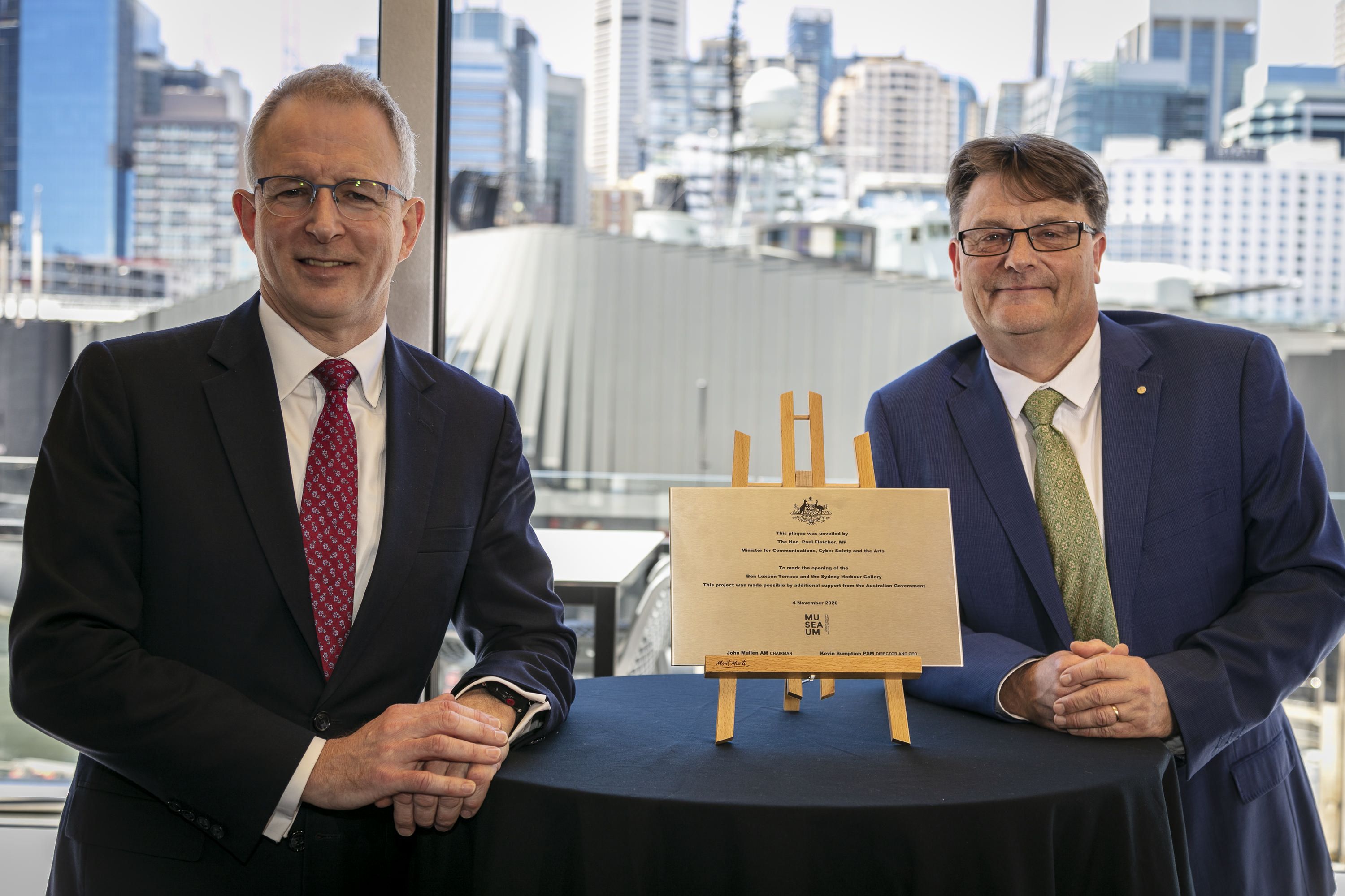 Photo of two men wearing suits, and standing next to a plaque.