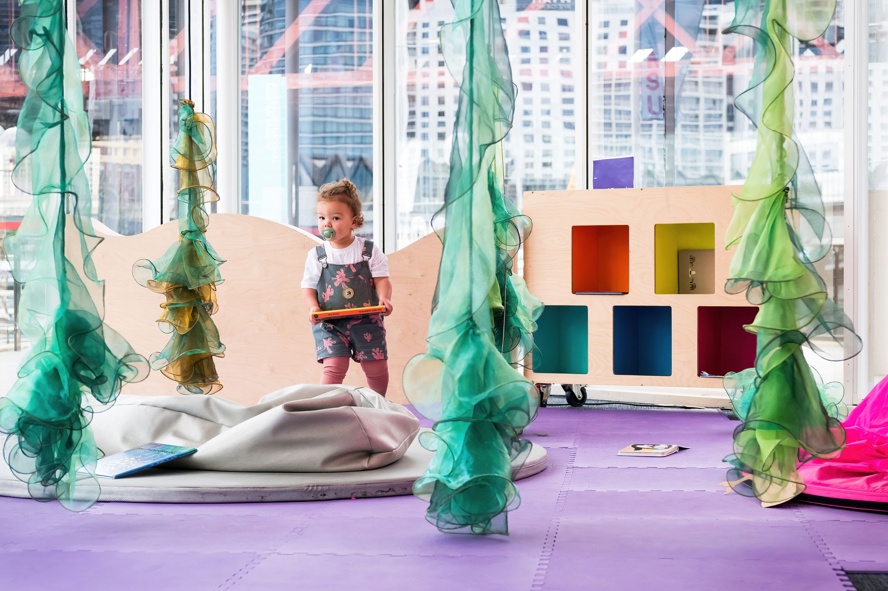 Photo of a toddler standing a holding a book in a bright, indoor space with colourful decorations.