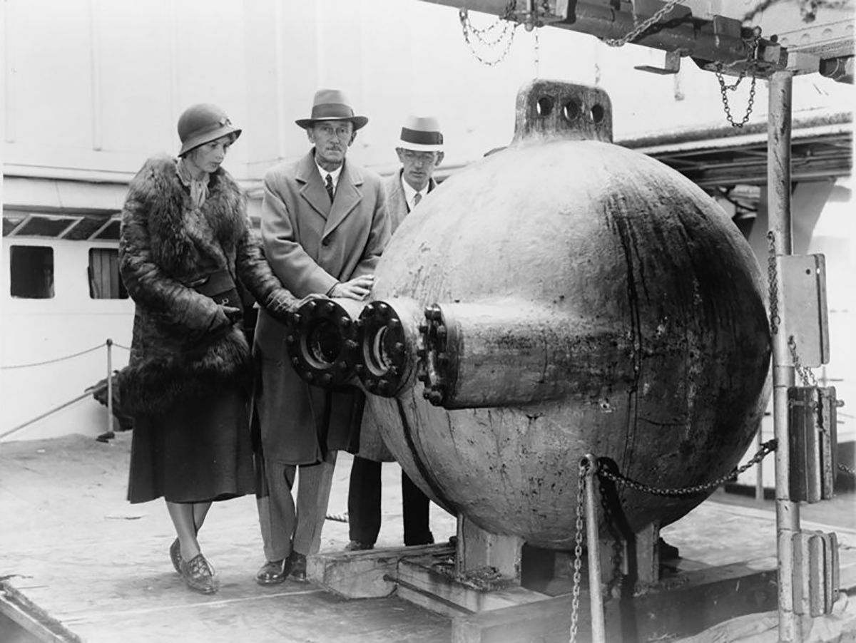 Black and white photo showing 3 well dressed people next to a round sphere.