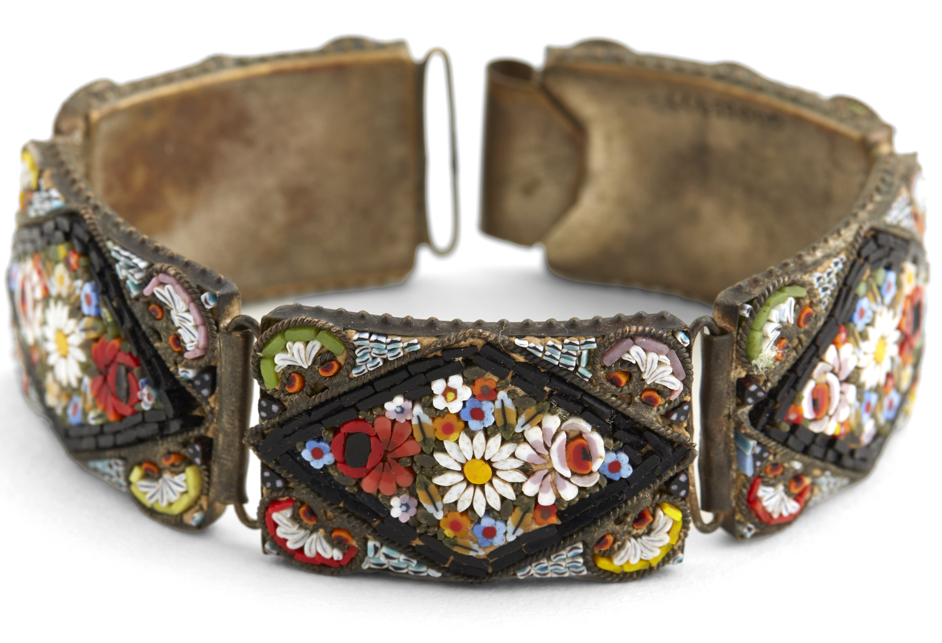 Italian bracelet ornamented with micro mosaic floral designs. The bracelet has five metallic rectangular panels decorated with small colourful tesserae arranged as flowers. Each panel has a diamond shaped area with a black outline and inside, floral arrangements that include a white flower with a yellow centre, a red flower with a black centre and a pink flower with white petals on the outside. There are smaller white, blue and orange flowers as well as leafs. Outside the diamond shaped area, there are semi