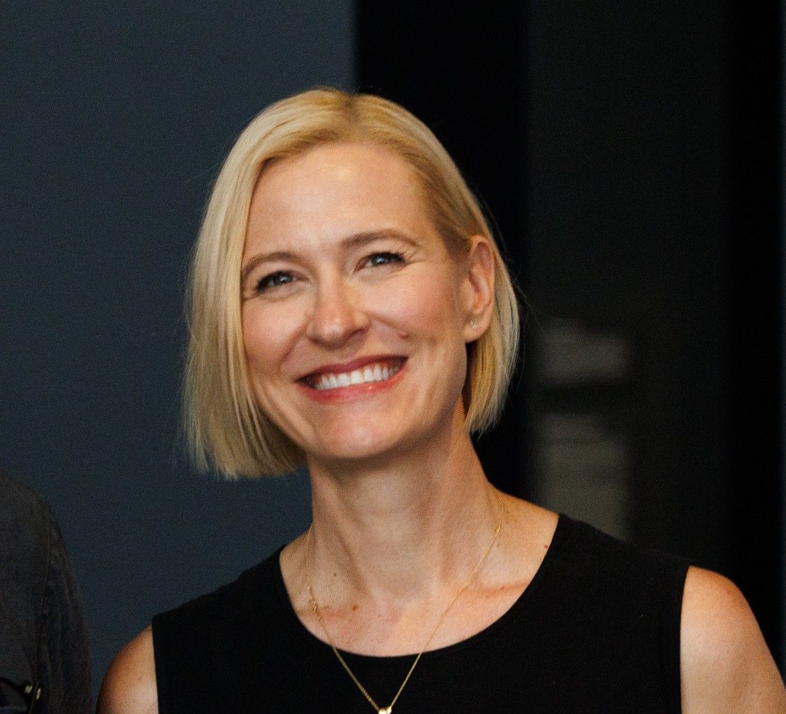 Photo of a woman smiting at the camera. She has short blonde hair and is wearing a black top.