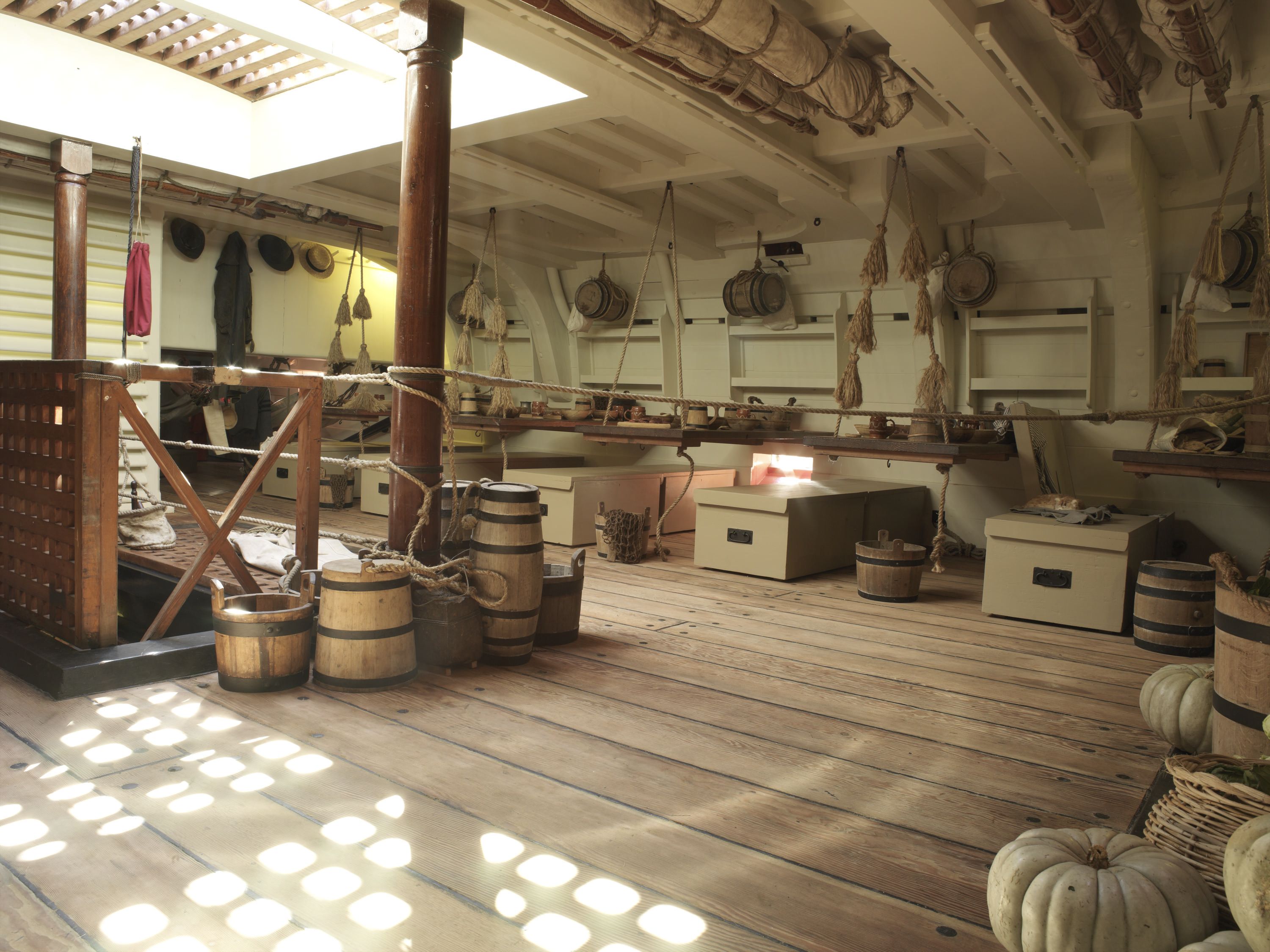 Photo showing the inside of a wooden tall ship, with sea chests, tables and barrels. 