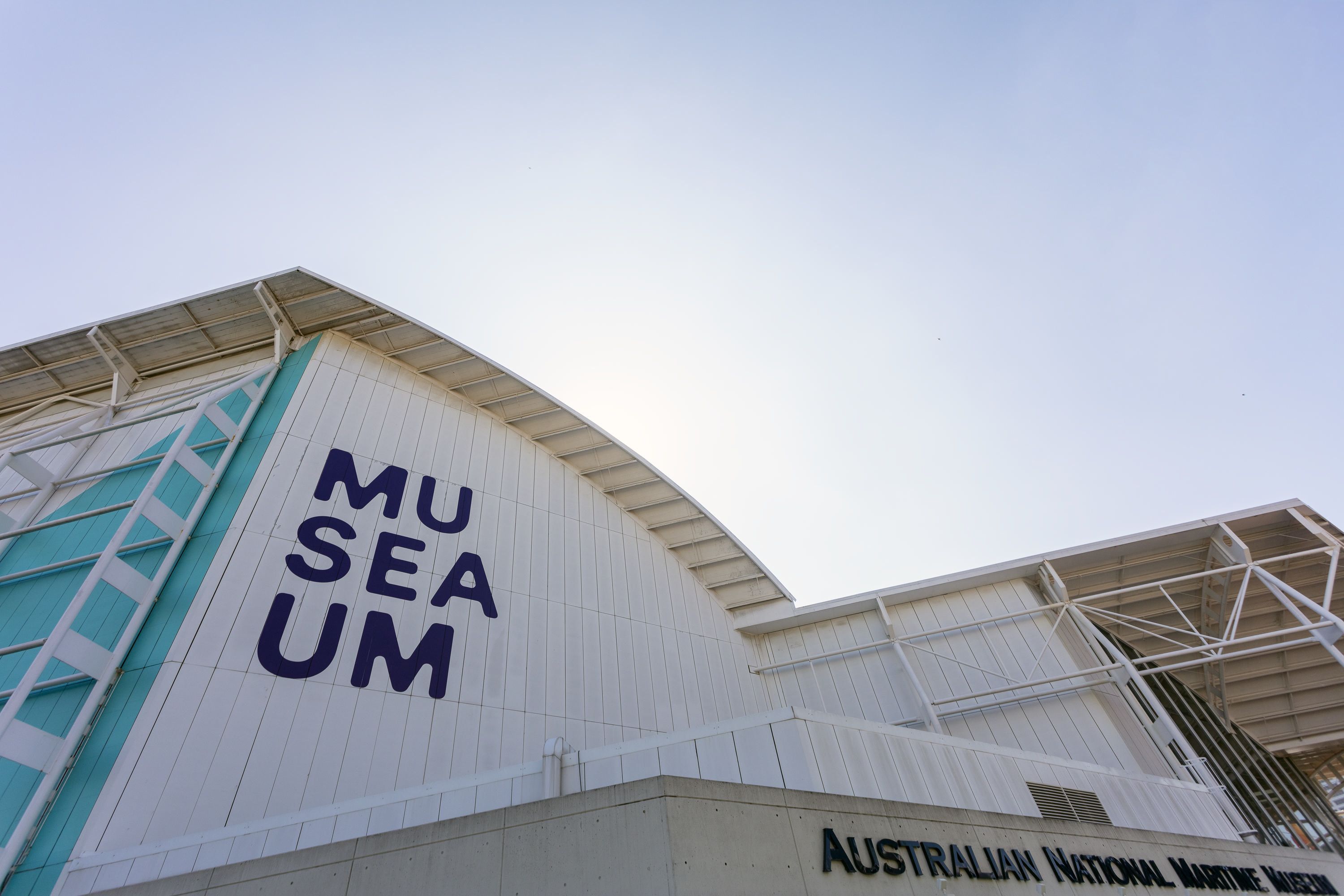 Photo of the museum building, with the logo on the white wall, and a blue sky behind. 
