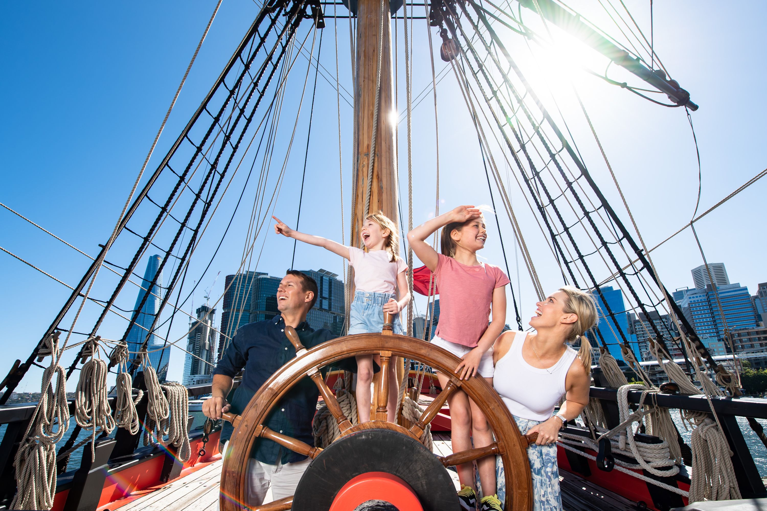 A family with two girls pose behind the ship's wheel on a wooden tall ship.
