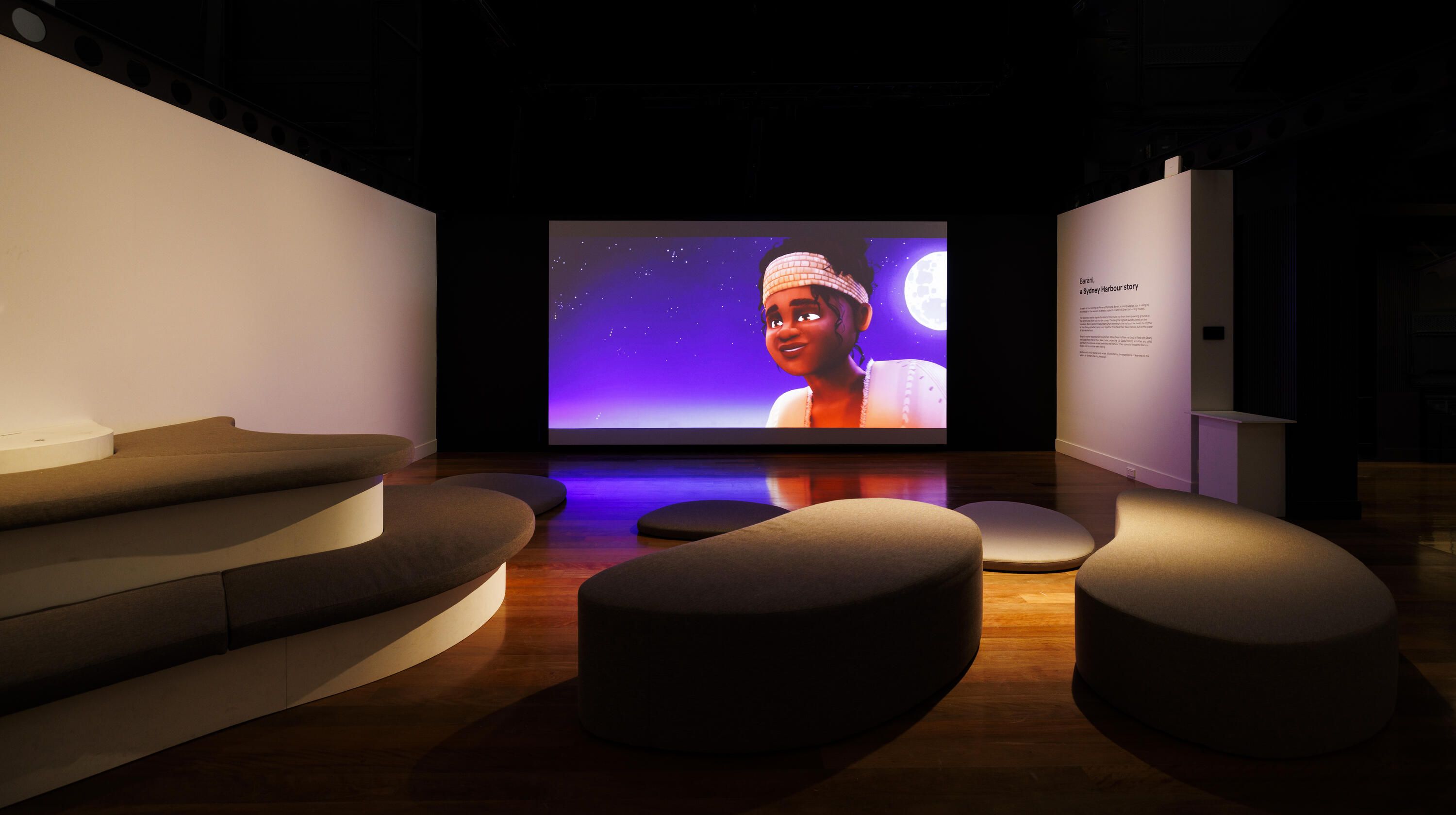 Photo of a museum exhibition space with curved soft seating and a large projection screen
