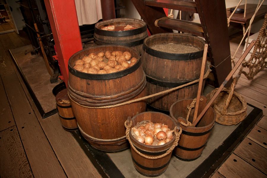 Photo taken of a museum display on a wooden tall ship. Wooden barrels contain food such as potatoes.