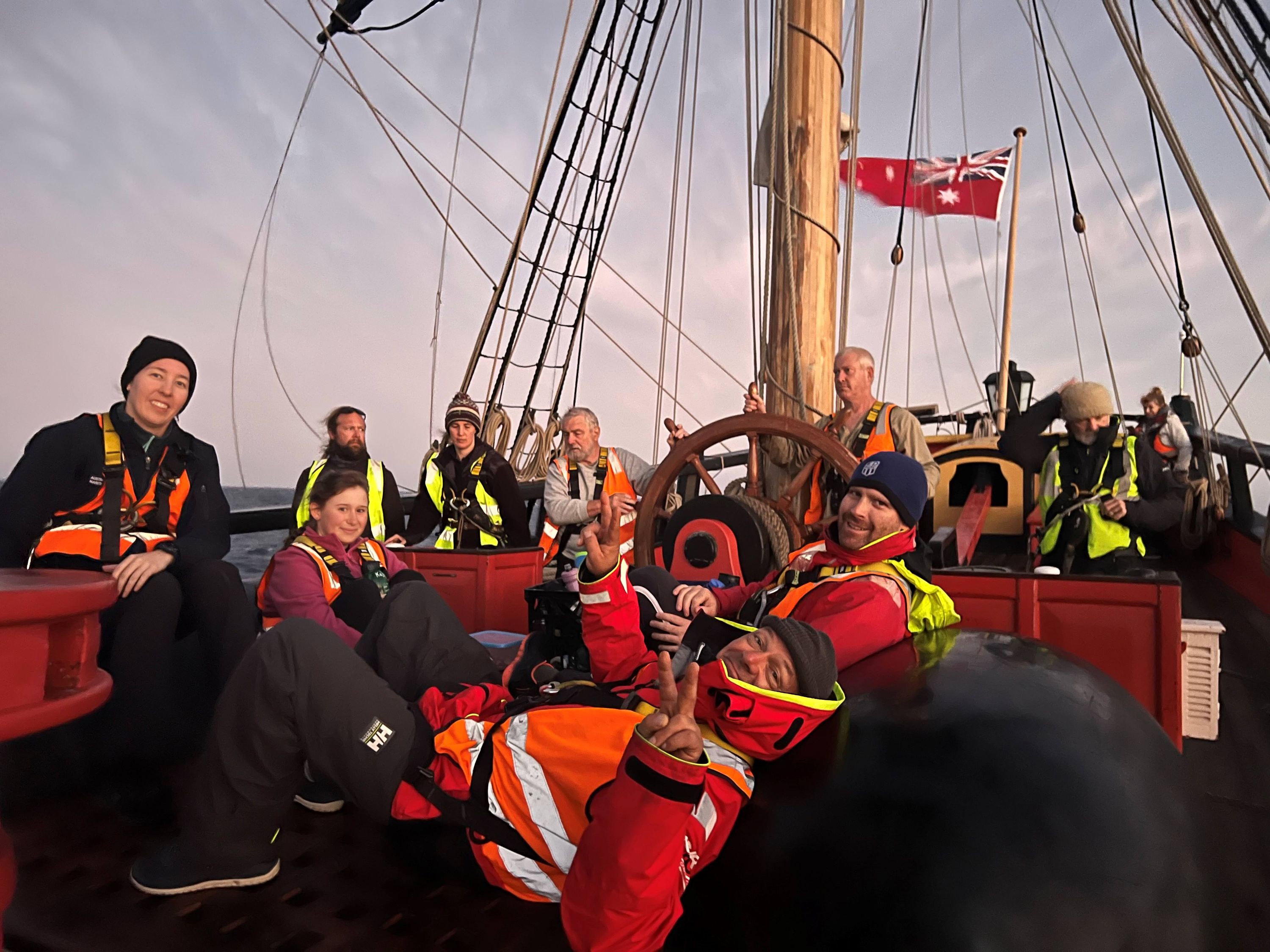 Group of people sitting on the deck of a tall ship wearing warm clothes, Fluro vests and climbing harnesses