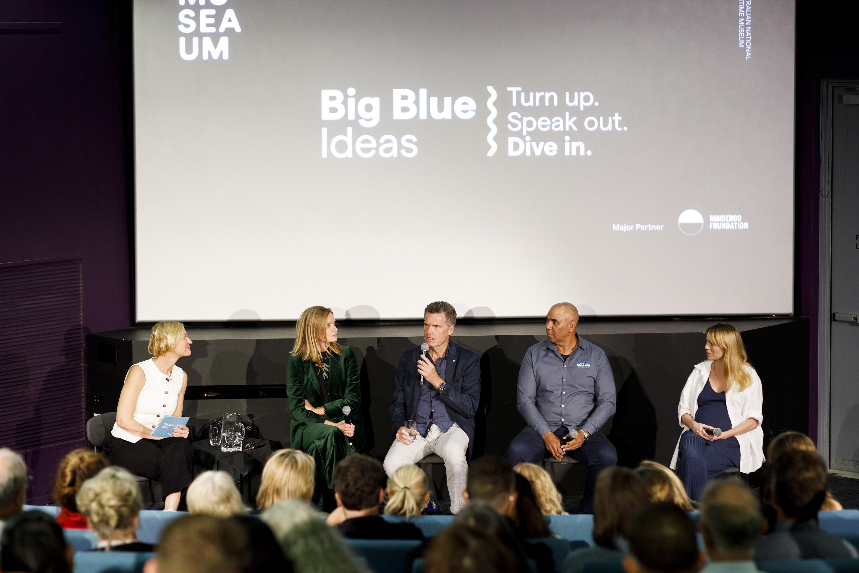 A photo showing a line of people seated beneath a screen at the front of a theatre. 