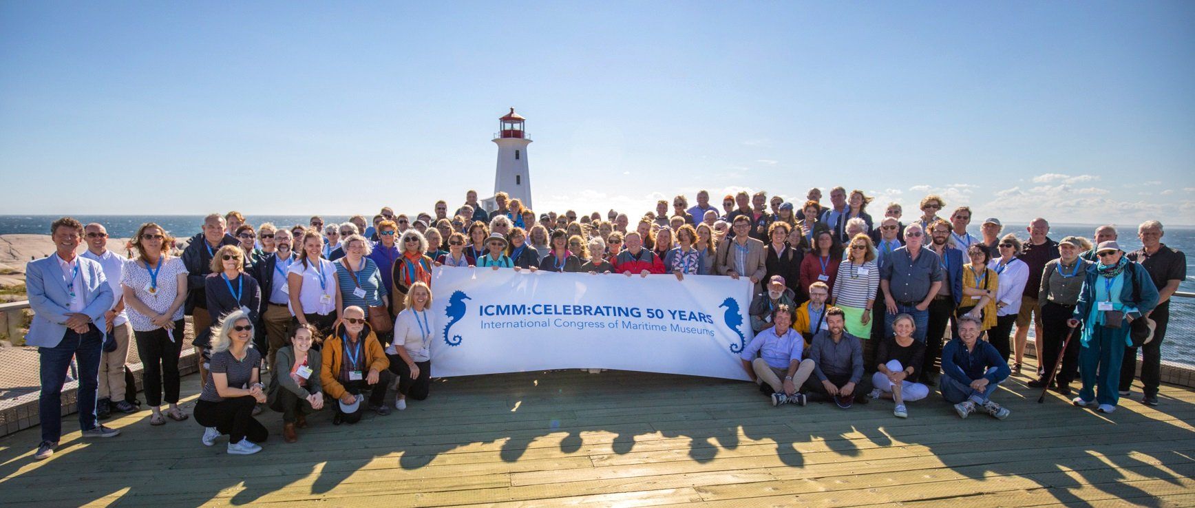 Photo of a large group of people holding a banner saying "ICMM: celebraiting 50 years" on a sunny day. There is a lighthouse in the background.