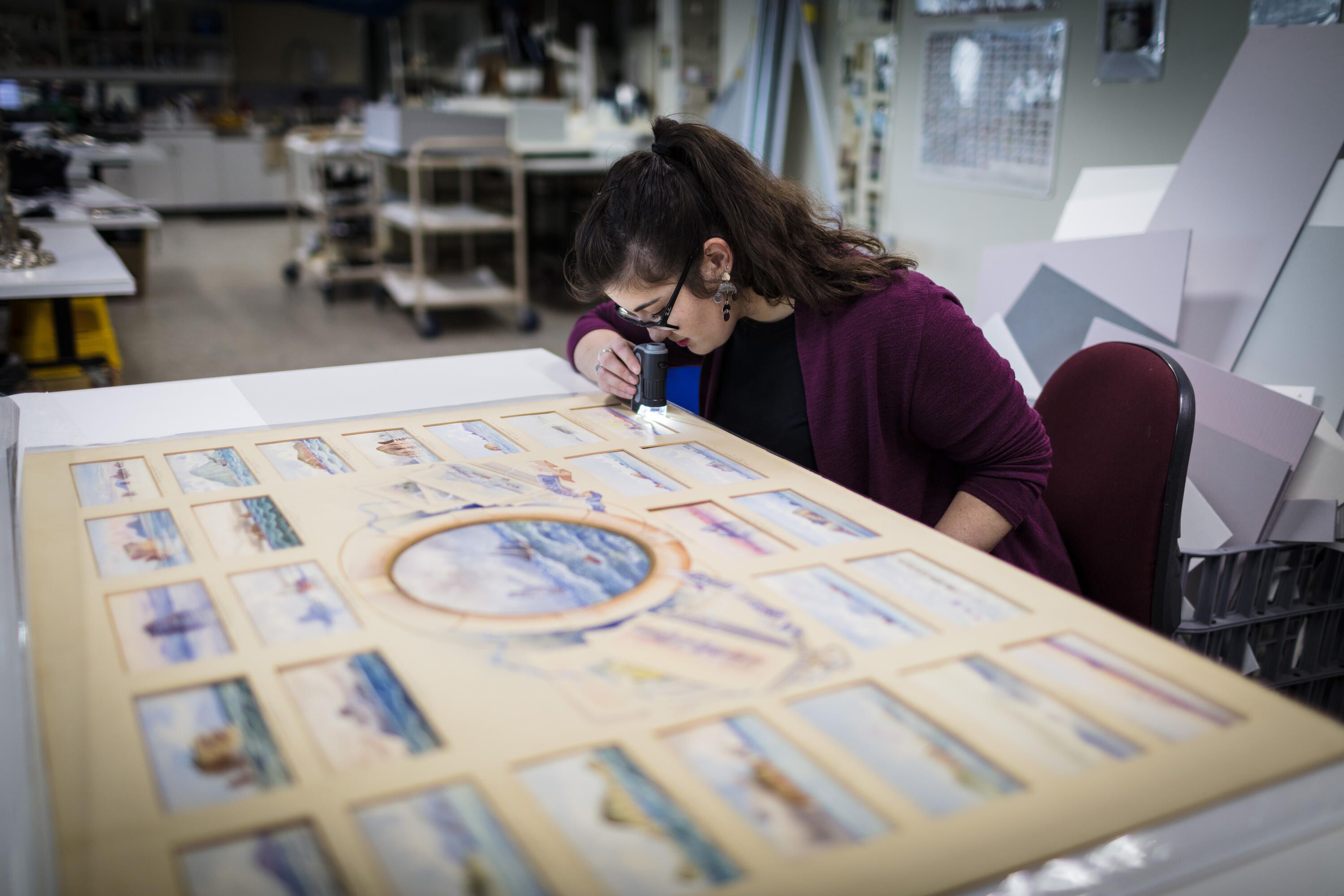 Photo of a woman with dark hair and a purple cardigan, using a magnifying glass to look at a watercolour artwork.