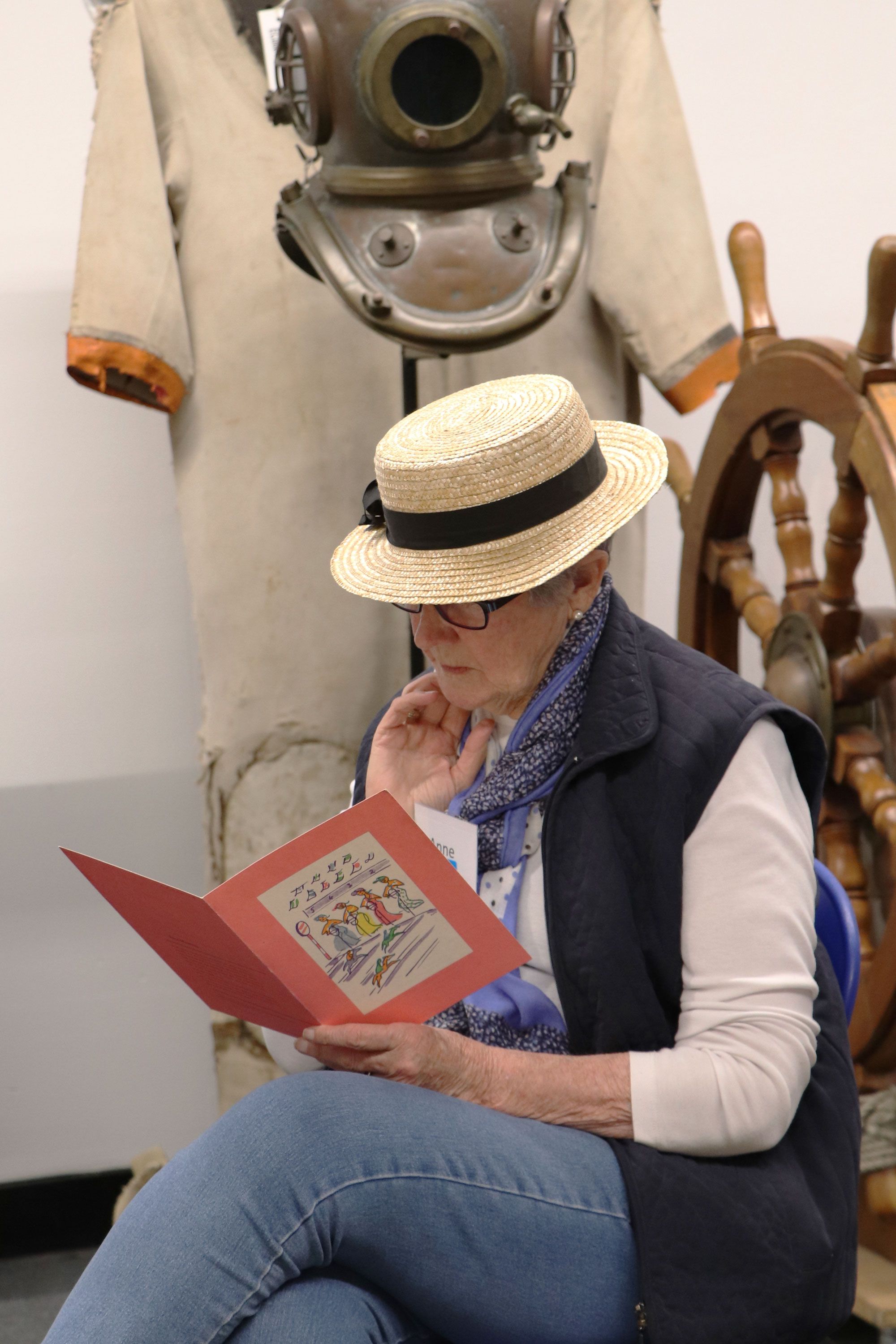 Lady wearing a straw hat and dark vest reading an old ship menue featuring a colourful painting on the cover