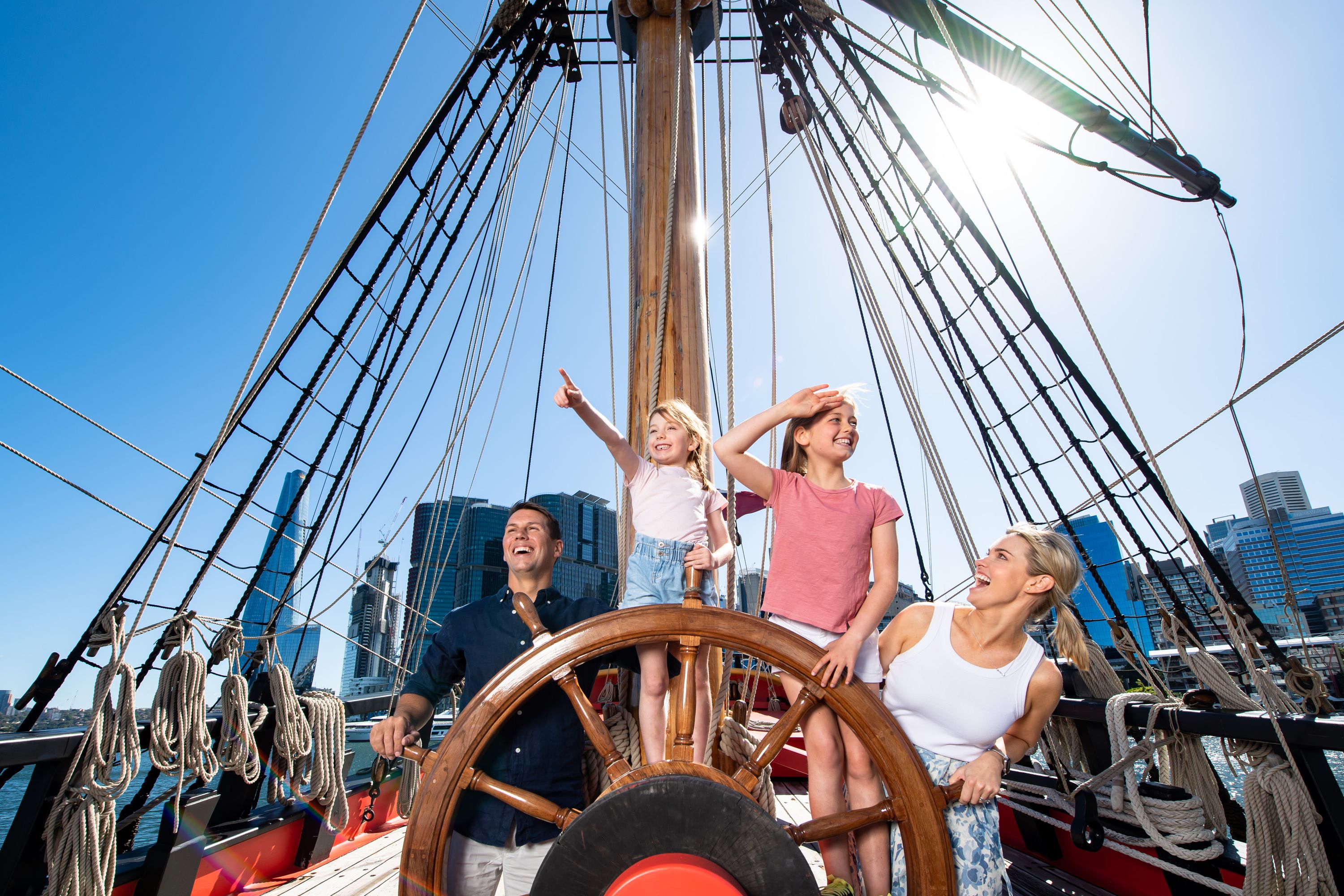 Photo showing a family with 2 girls standing behind the wheel of a wooden tall ship, smiling and pointing.