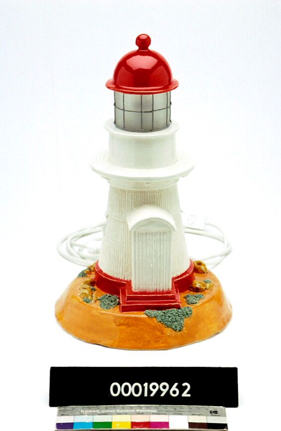This ceramic lamp is based on the Cooktown Lighthouse in Queensland. It was a red glazed ceramic roof. White glaze has been used for the tower of the lighthouse and fashioned to resemble corrugated iron. The design also includes three barred windows and an entrance hall. The foundation of the lighthouse and the stairs leading to the door are treated with a red glaze. The lighthouse sits on a raised hillock with rocks and vegetation, respectively treated with green and brown glazes.