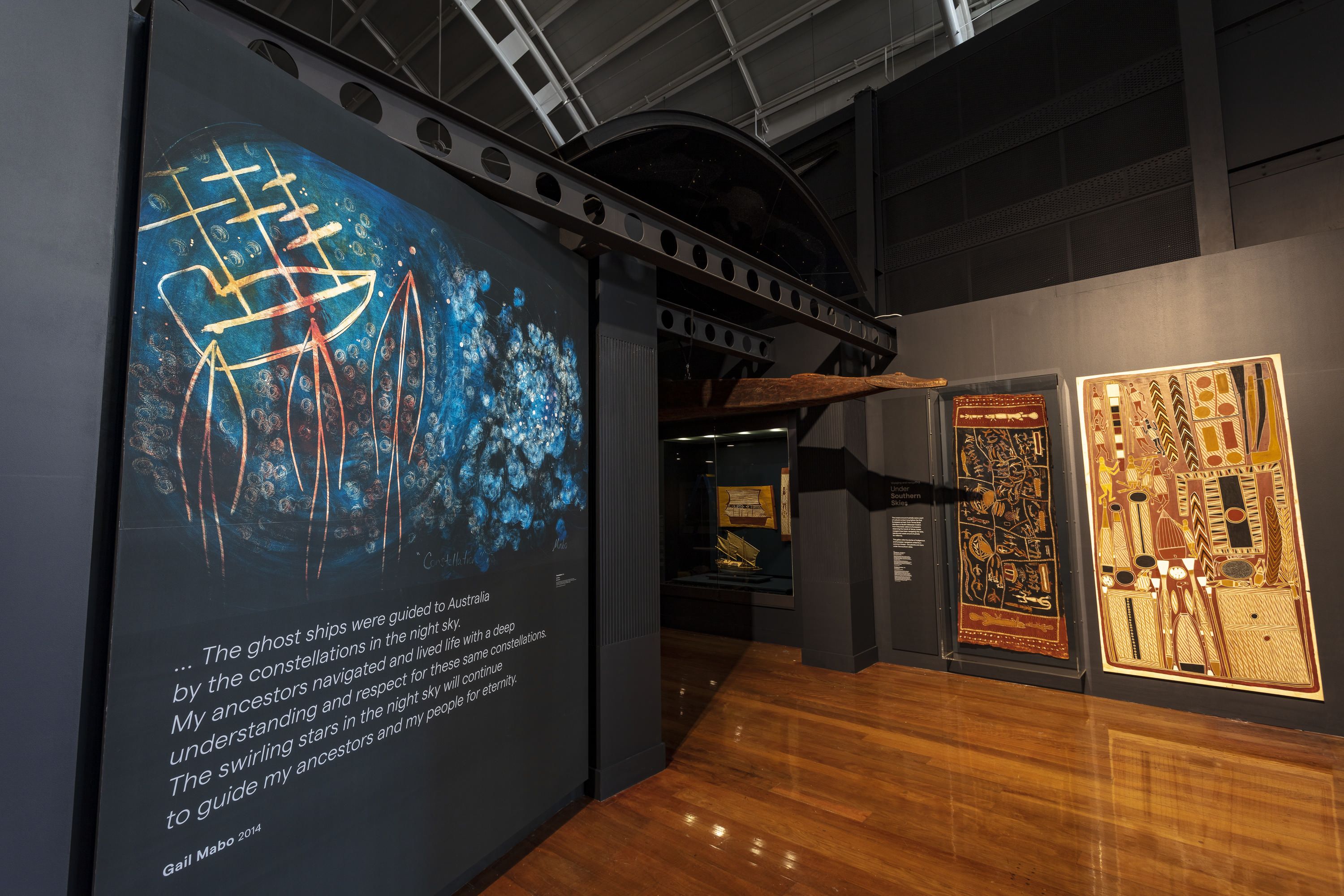 Photo showing the entrance to a museum gallery, there is a large print of a painting on the left and 2 bark paintings on the right. 